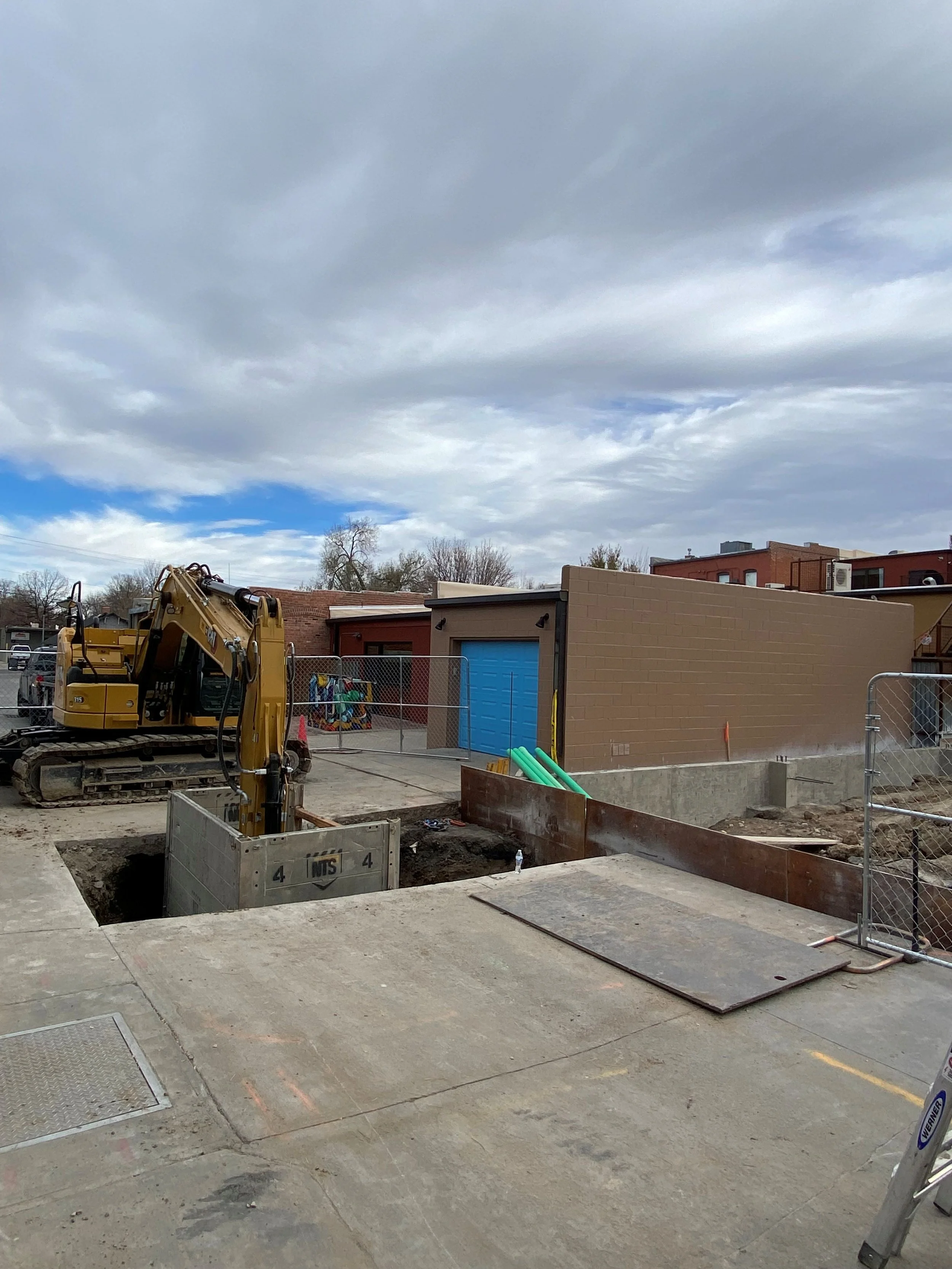 Construction site with a small excavator, open excavation, safety barrier, and nearby neighboring buildings with a blue garage door and brick wall under a cloudy sky.