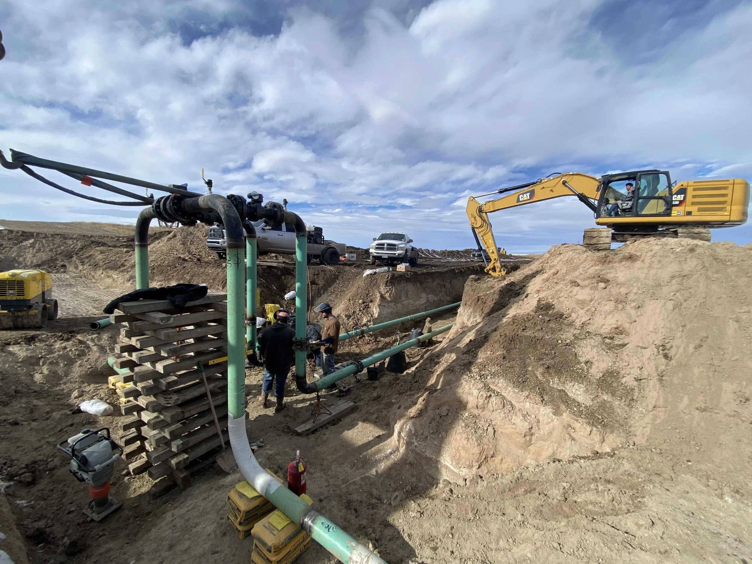 Construction site with workers installing large green pipes, an excavator on a pile of dirt, and several vehicles in the background under a partly cloudy sky.