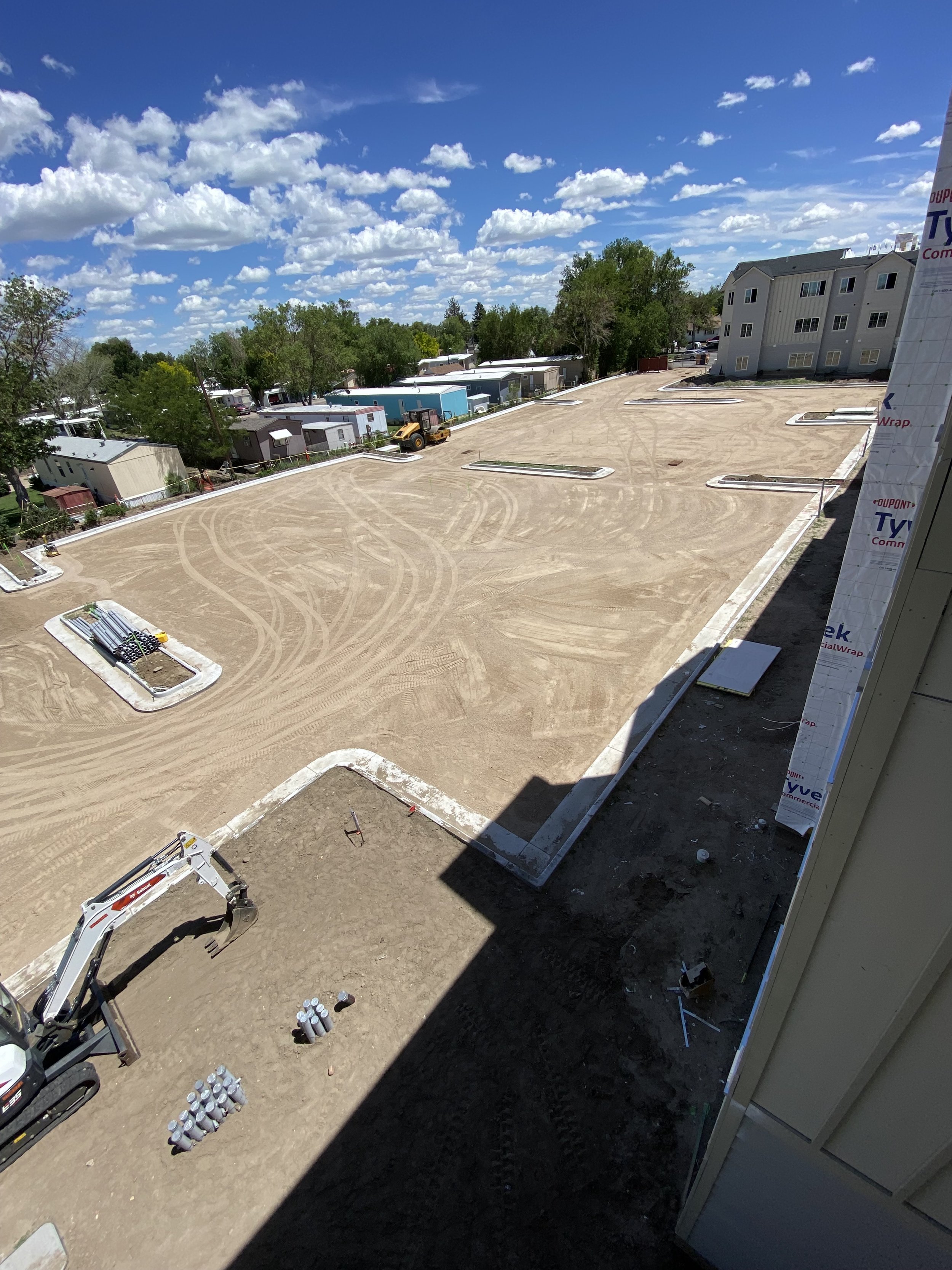 Construction site with a cleared dirt lot, construction materials, and a small excavator, under a partly cloudy blue sky, with residential buildings in the background.