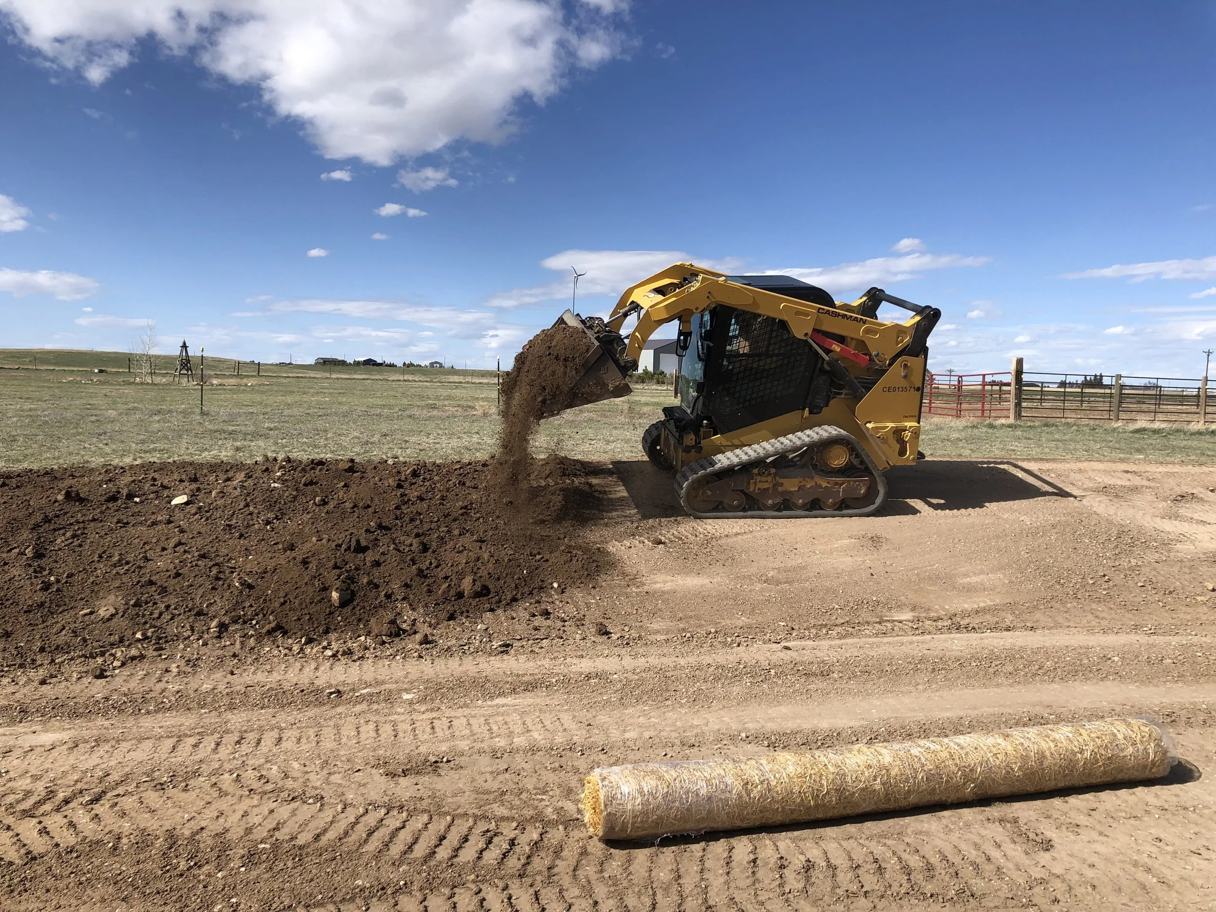 A yellow skidsteer moving dirt on a construction site with a clear blue sky and clouds, fencing, and rural landscape in the background.