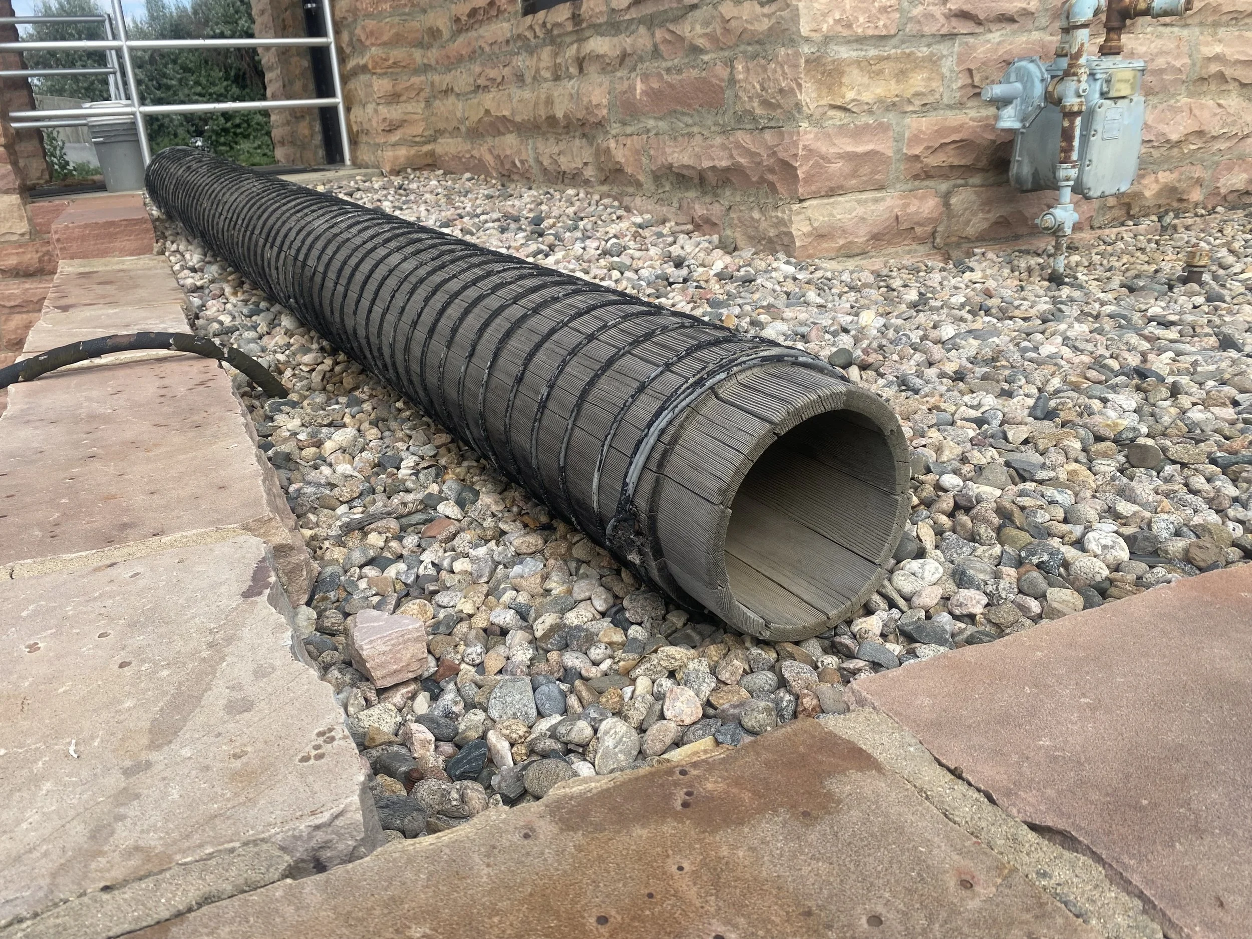 A large, black, flexible drainage pipe lying on a gravel surface next to a brick wall and stone pavers, with a metal water meter and pipes attached to the wall in the background.