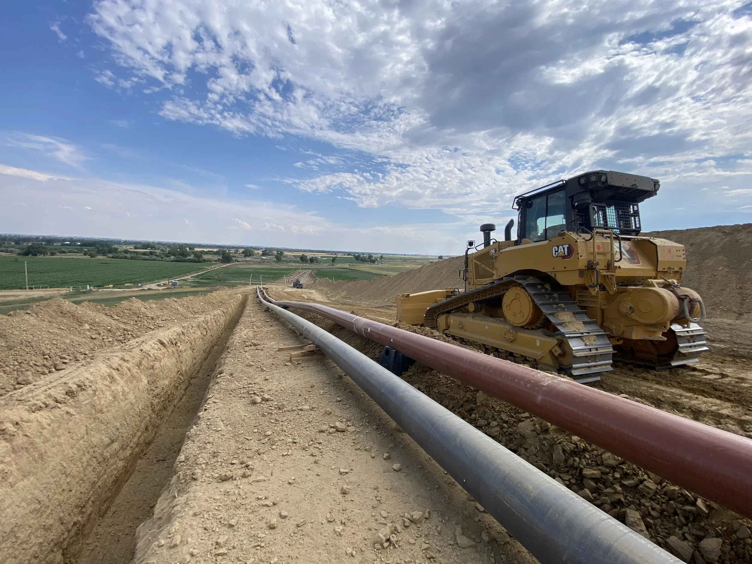 A construction site with a CAT bulldozer on dirt, laying pipes in a long trench, with farmland and a blue sky with clouds in the background.