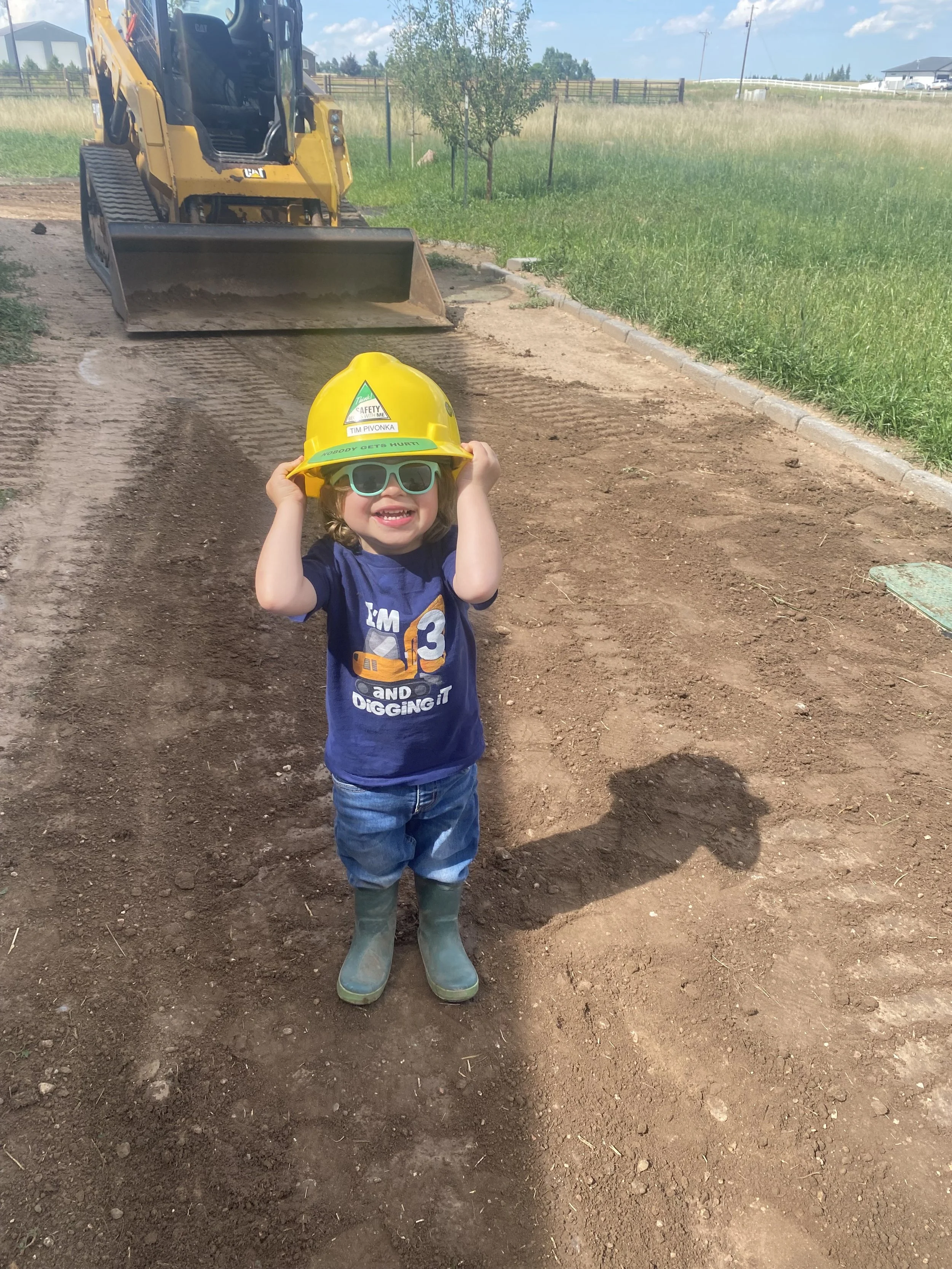 Young boy at a construction site wearing a yellow safety helmet, blue sunglasses, rubber boots, and a navy T-shirt, smiling and adjusting his helmet, with a skidsteer and green grass in the background.