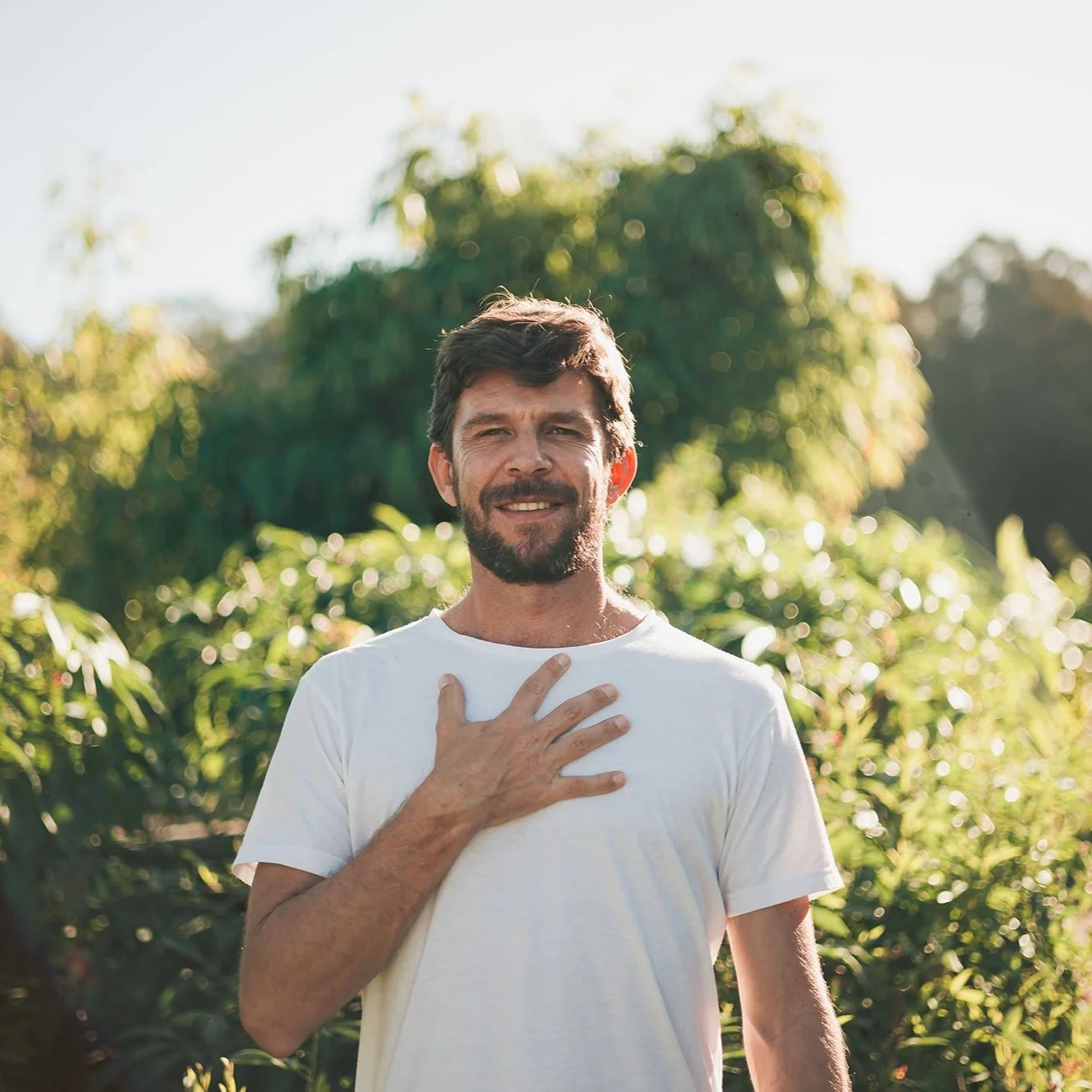 A man with a beard and brown hair, wearing a white t-shirt, standing outdoors with greenery in the background. He has his right hand on his chest and is smiling, with sunlight illuminating the scene.