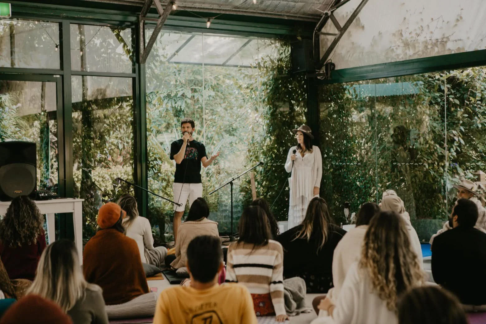 Two people stand on stage speaking into microphones in front of an audience in a glass-walled indoor space with green plants outside the windows.