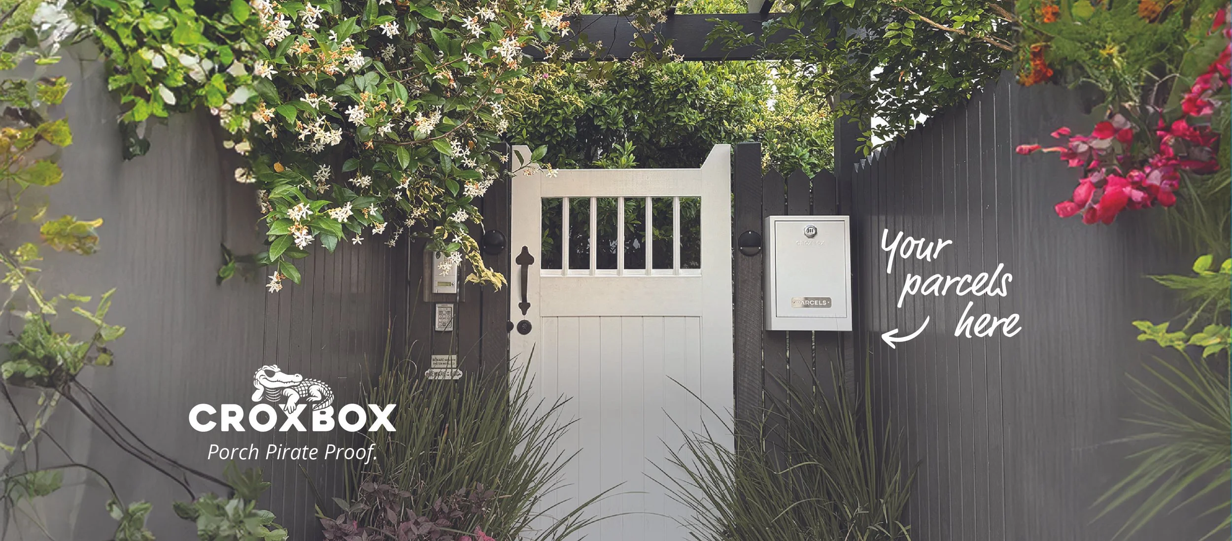 A grey fence with a white gate surrounded by green and colorful flowering plants. There is a mailbox or metal parcelbox on the right side of the gate with the text "Your parcels here" written next to it, indicating a safe drop area for deliveries.