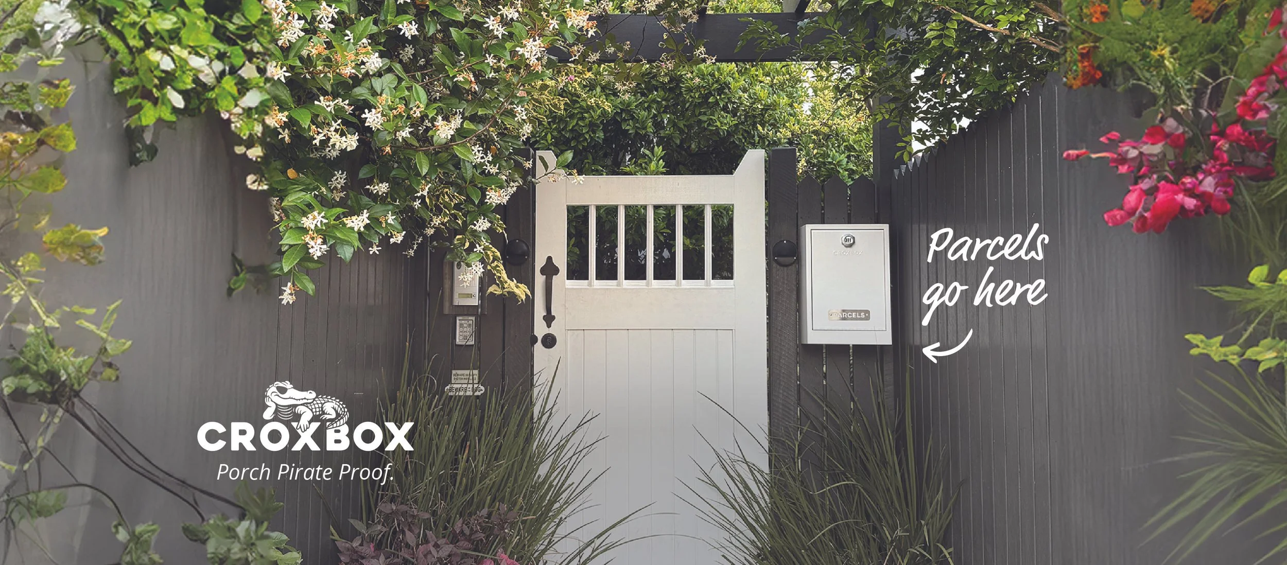 A white metal parcelbox with a CroxBox logo is attached to a dark grey fence in front of a garden with lush green plants, with text indicating parcels go here for easy delivery and a CrocBox logo with a crocodile wearing an eye patch.