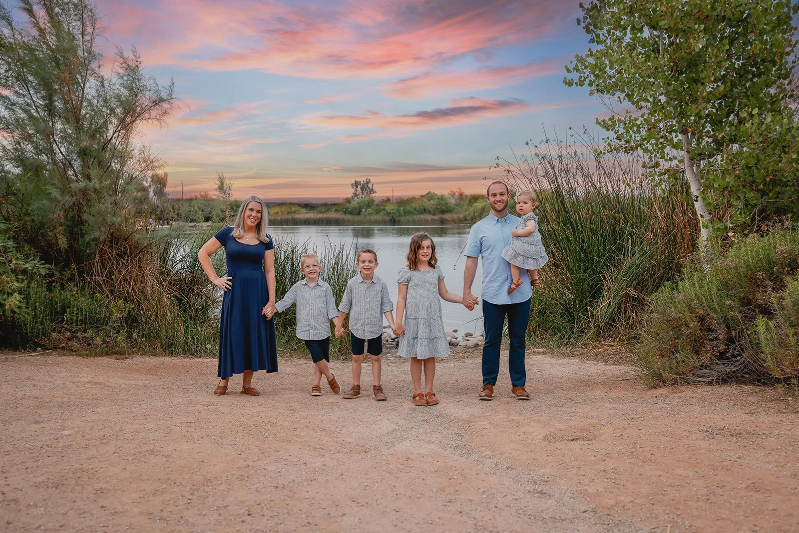 A family of six posing outdoors near a lake at sunset, with trees and bushes in the background.