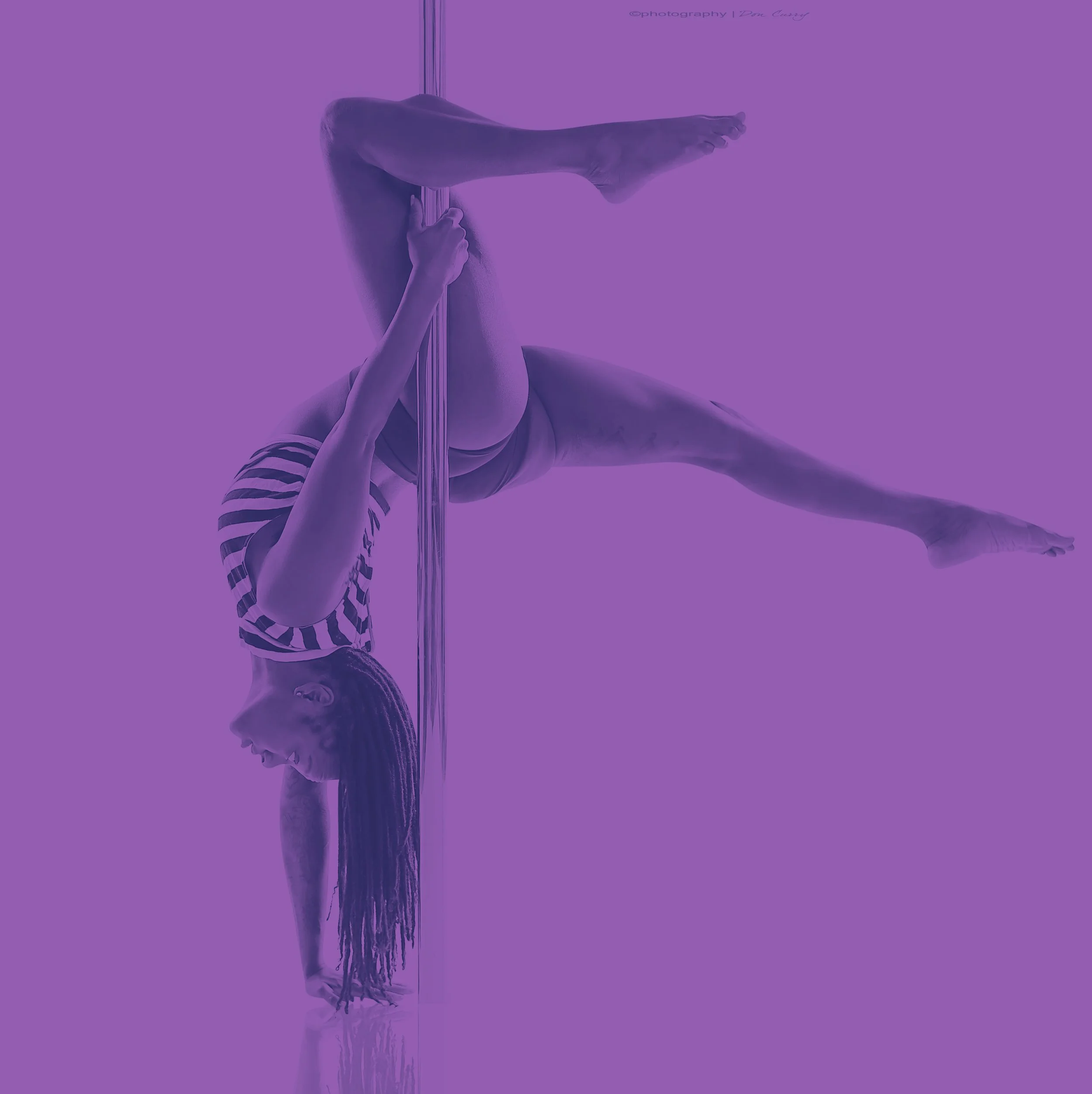 Person performing a pole dance move, hanging upside down and holding the pole with their legs, with their hands gripping the pole. The image has a purple filter overlay.