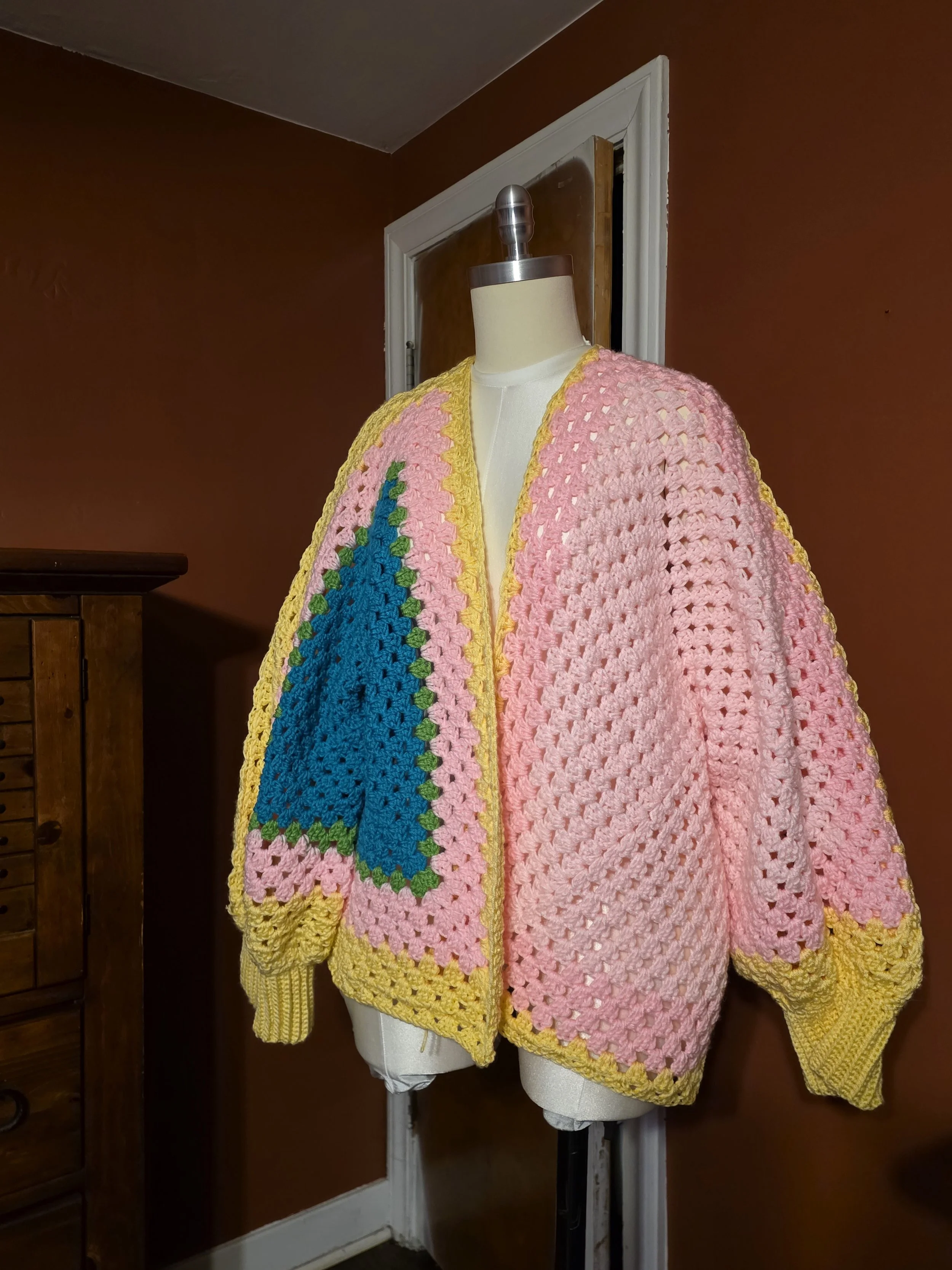 A mannequin wearing a colorful, hand-crocheted oversized jacket with pink, blue, green, and yellow sections, displayed indoors with a brown wall and wooden furniture in the background.