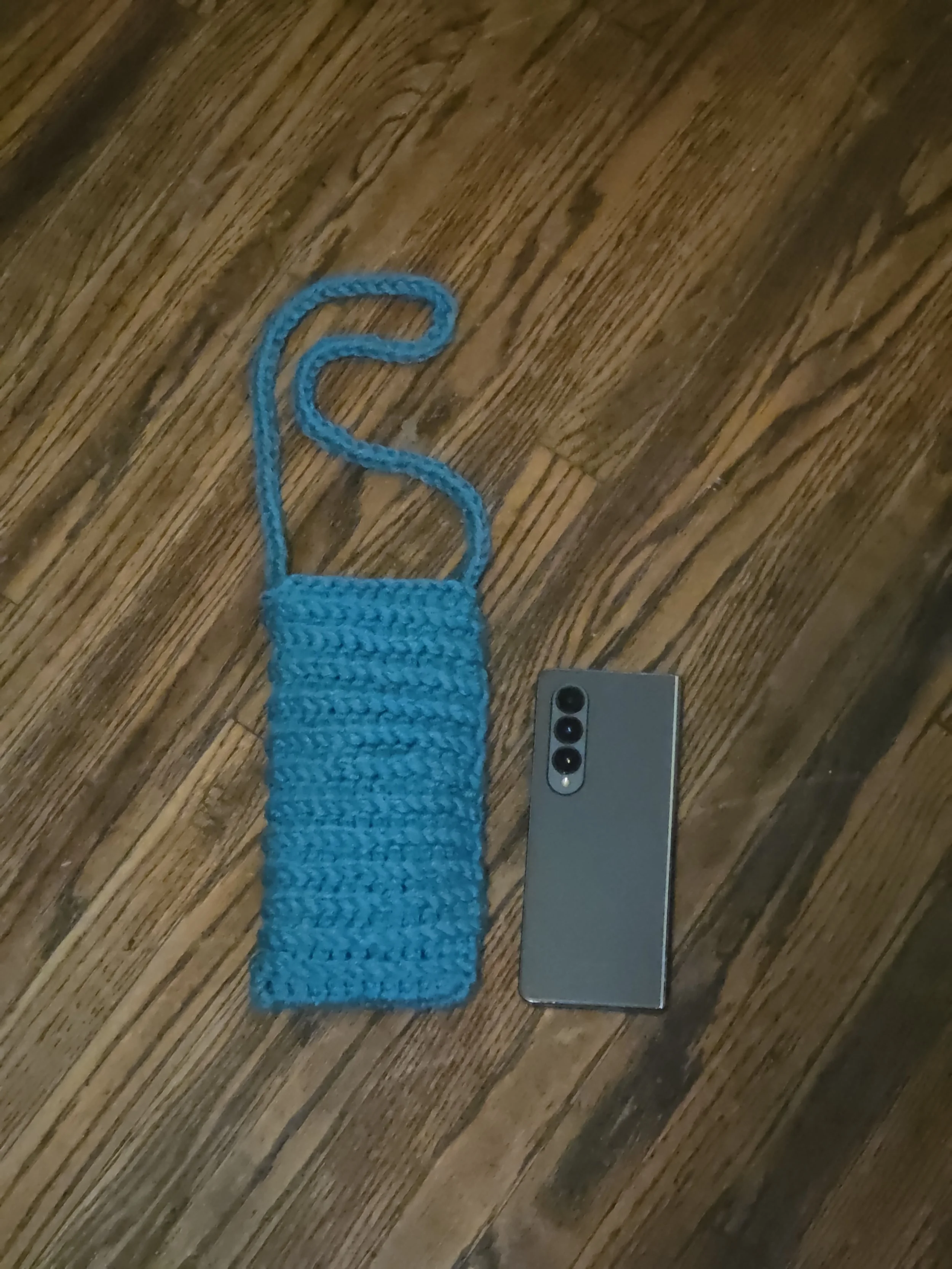 A blue crocheted phone holder with a strap and a silver phone with three camera lenses resting on a wooden floor.