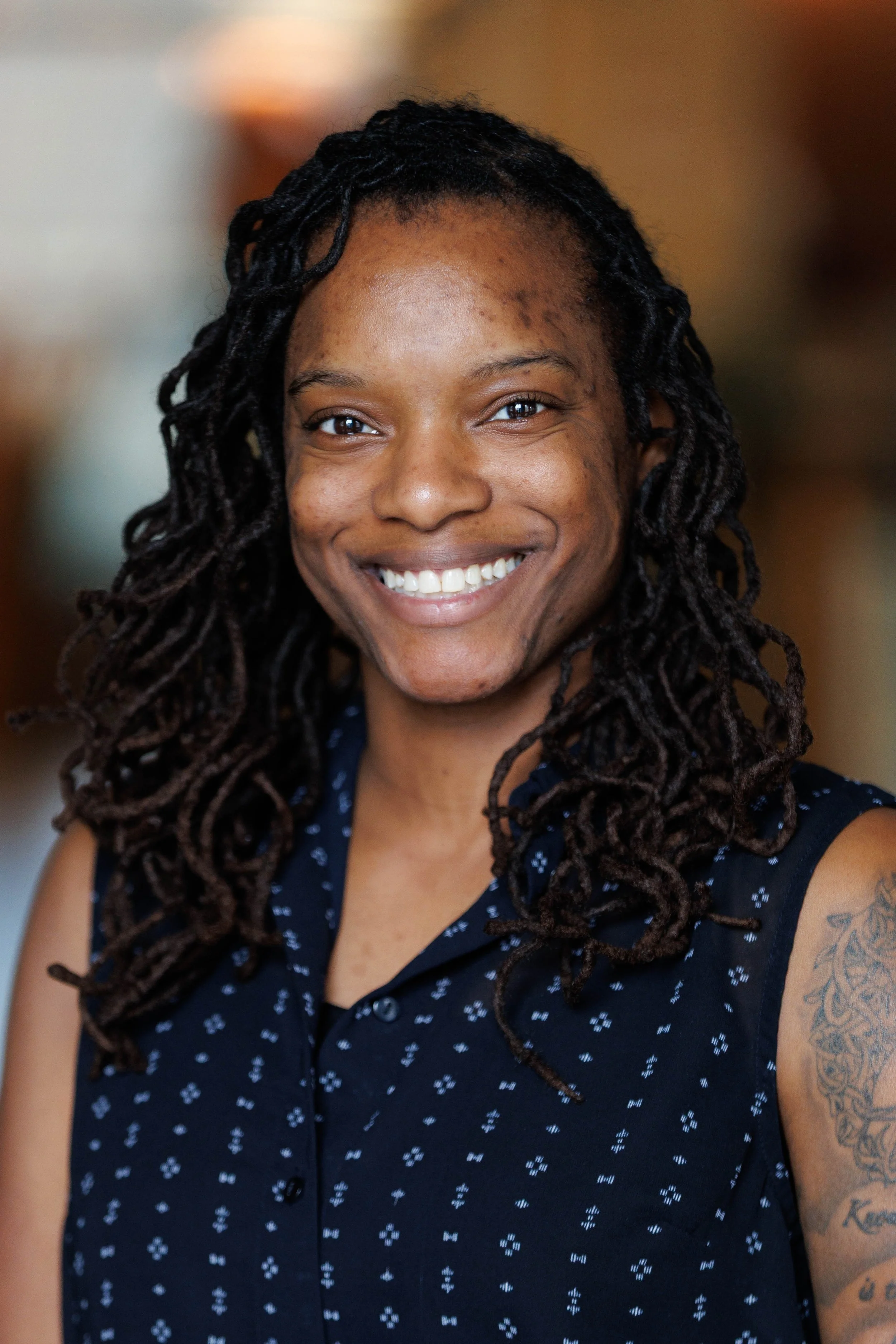 Smiling woman with shoulder-length dreadlocks wearing a sleeveless navy blue top with white floral pattern, in an indoor setting.