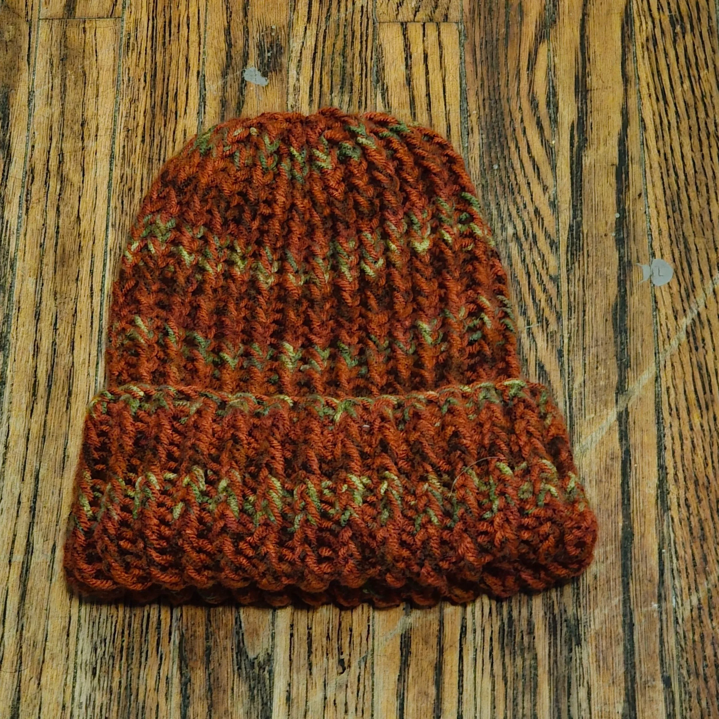 Knitted beanie with orange, green, and brown yarn placed on a wooden surface.