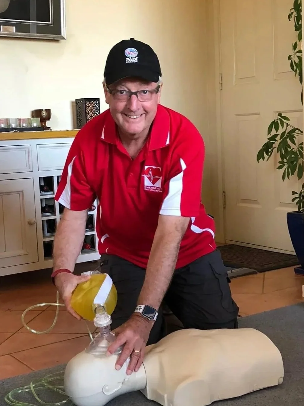 A man in a red emergency responder shirt and cap practicing CPR on a mannequin.