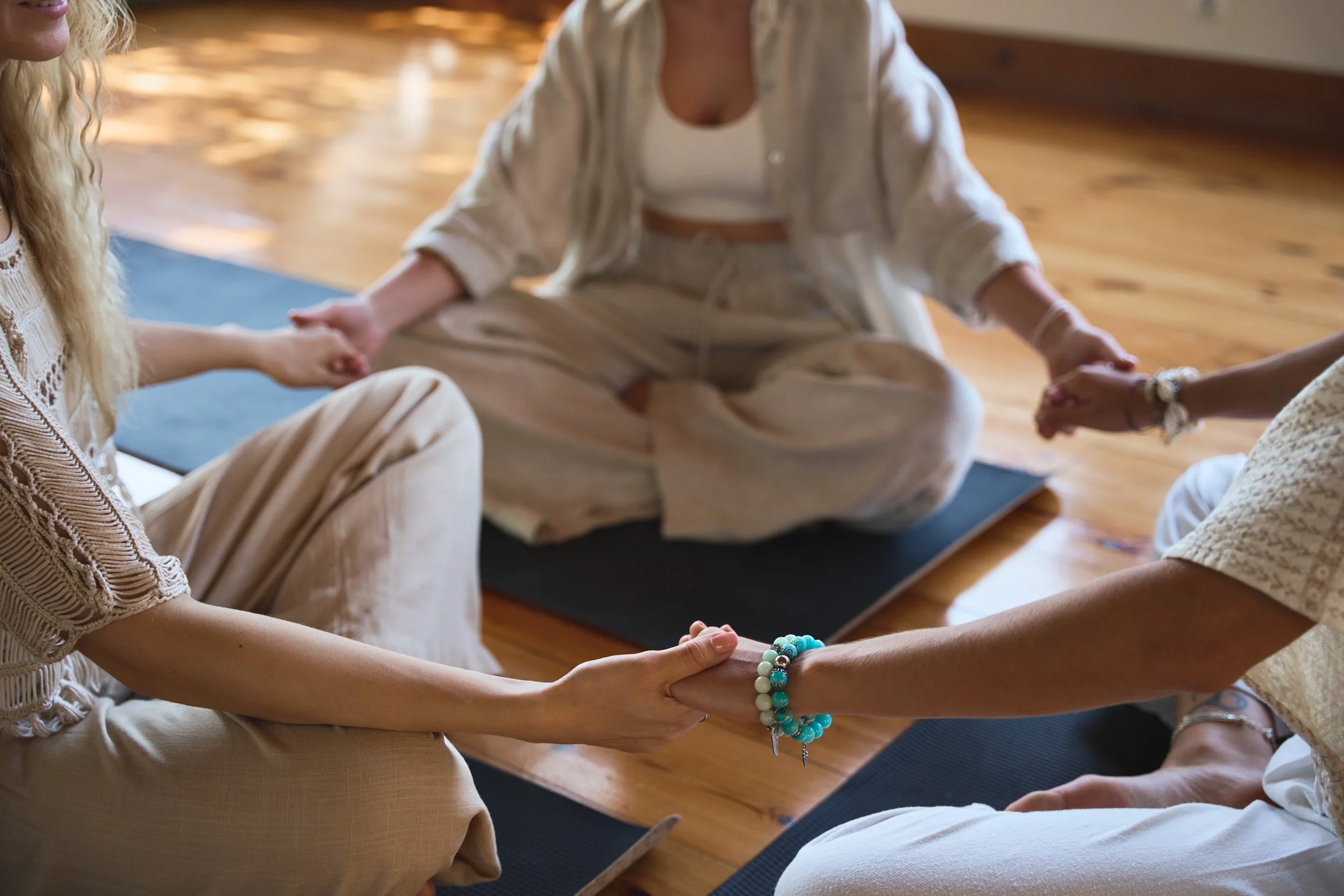 Three people sitting cross-legged on yoga mats holding hands in a circle during a meditation session in a room with wooden floors.