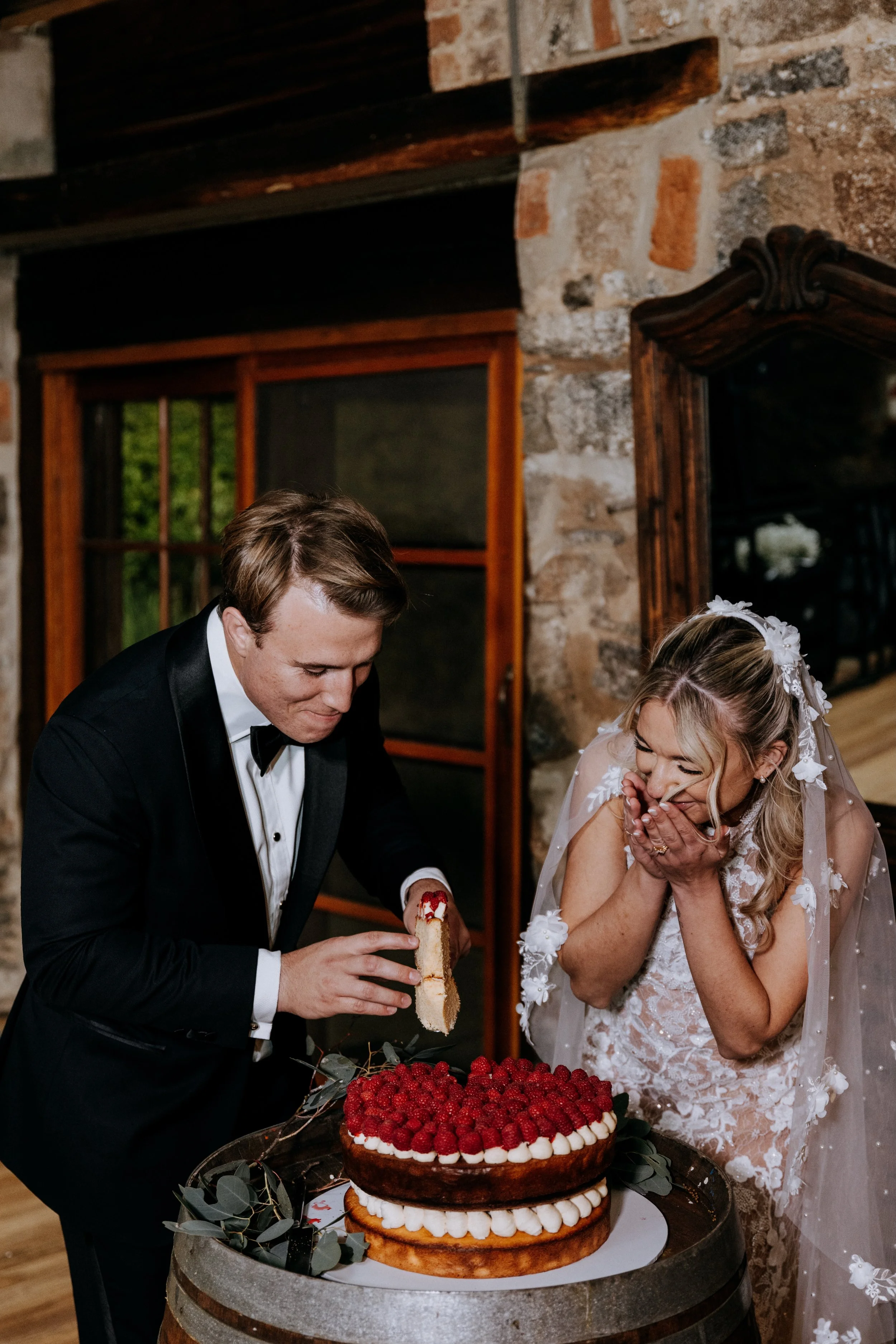 A newlywed couple at a wedding celebration, cutting a large wedding cake decorated with raspberries, strawberries, and cream, with a rustic brick wall and windows in the background.