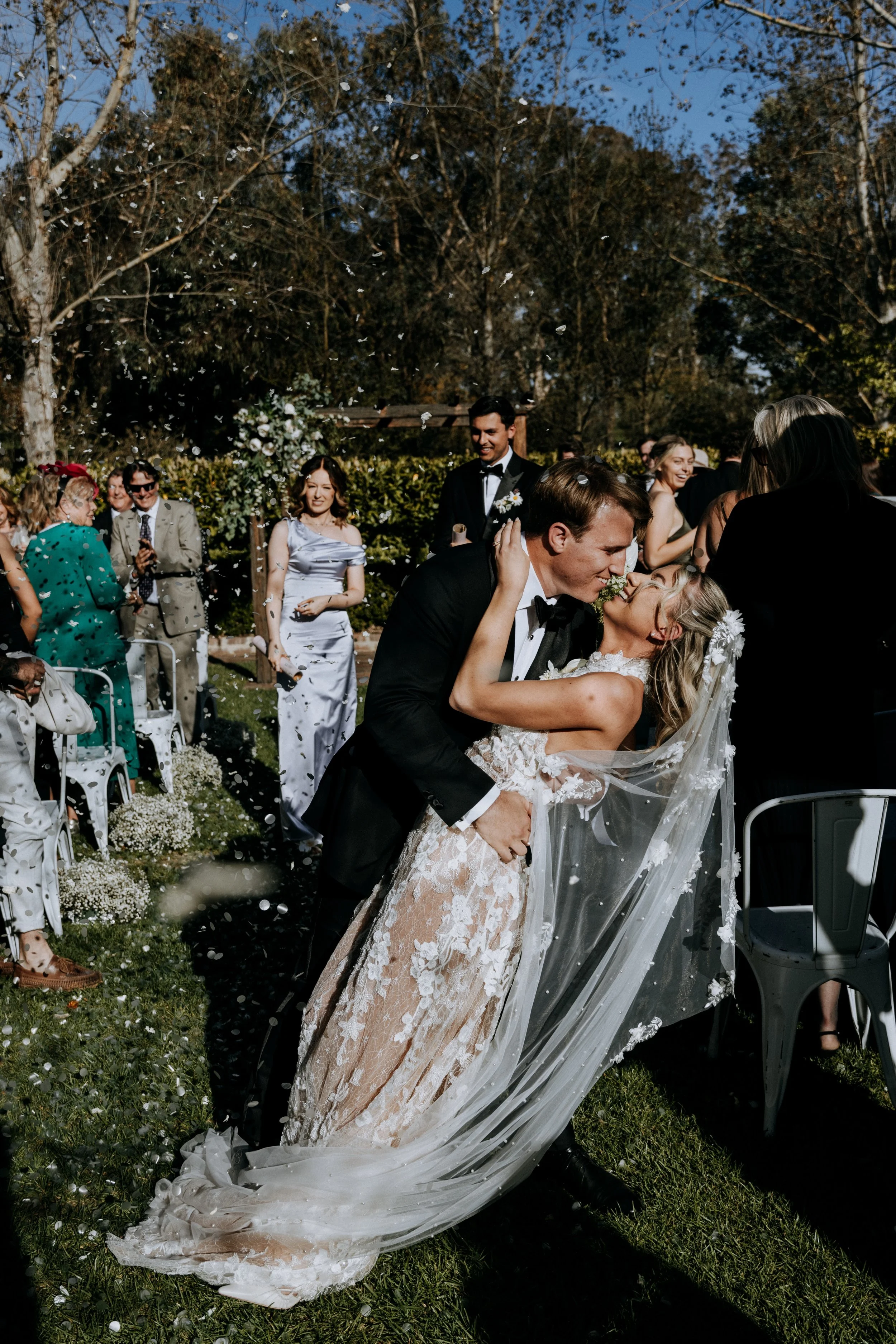 Couple kissing during outdoor wedding ceremony, surrounded by guests and falling flower petals on grass with chairs and trees in background.