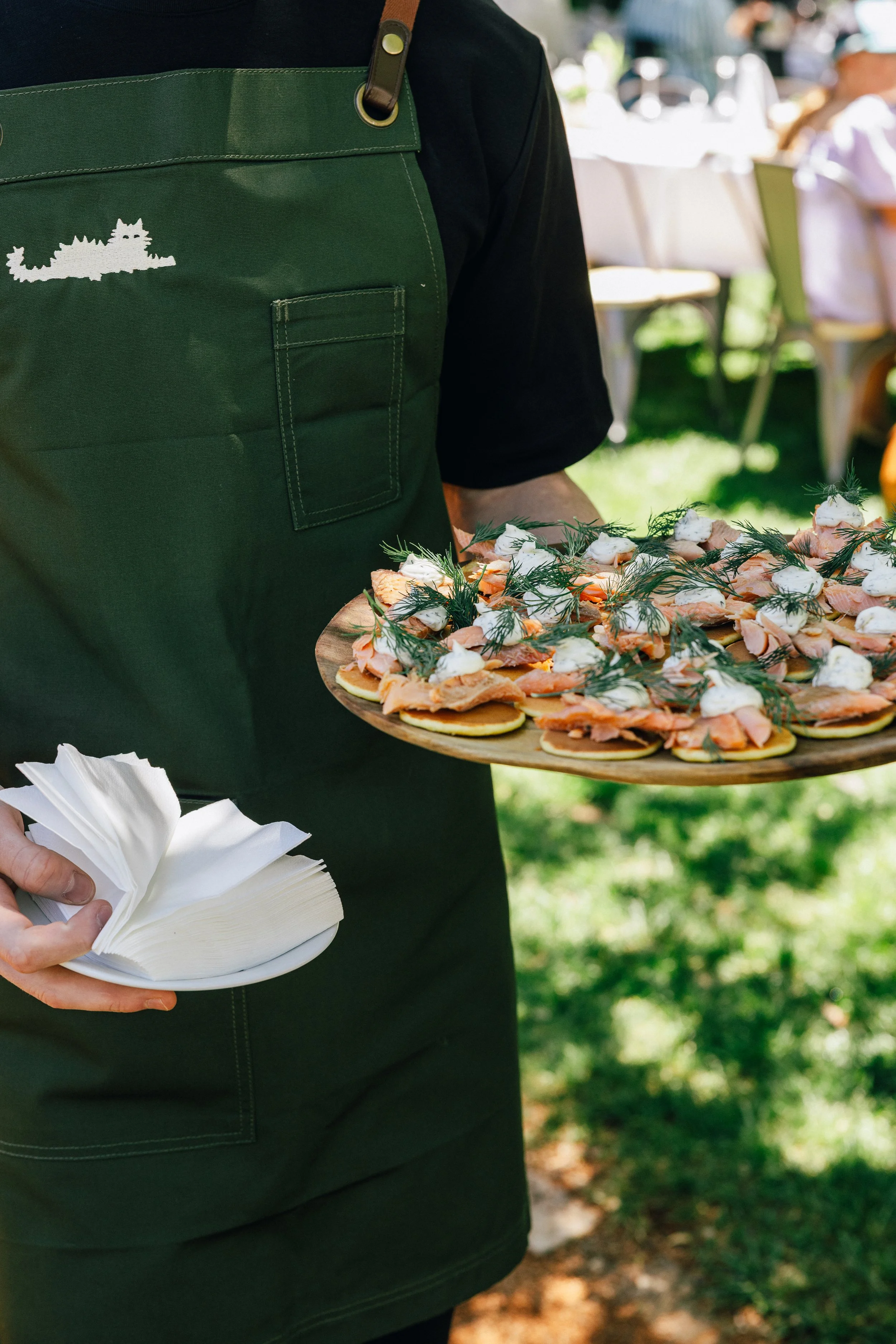 A server wearing a black shirt and green apron holding a white plate with small appetizers topped with herbs, and a napkin.
