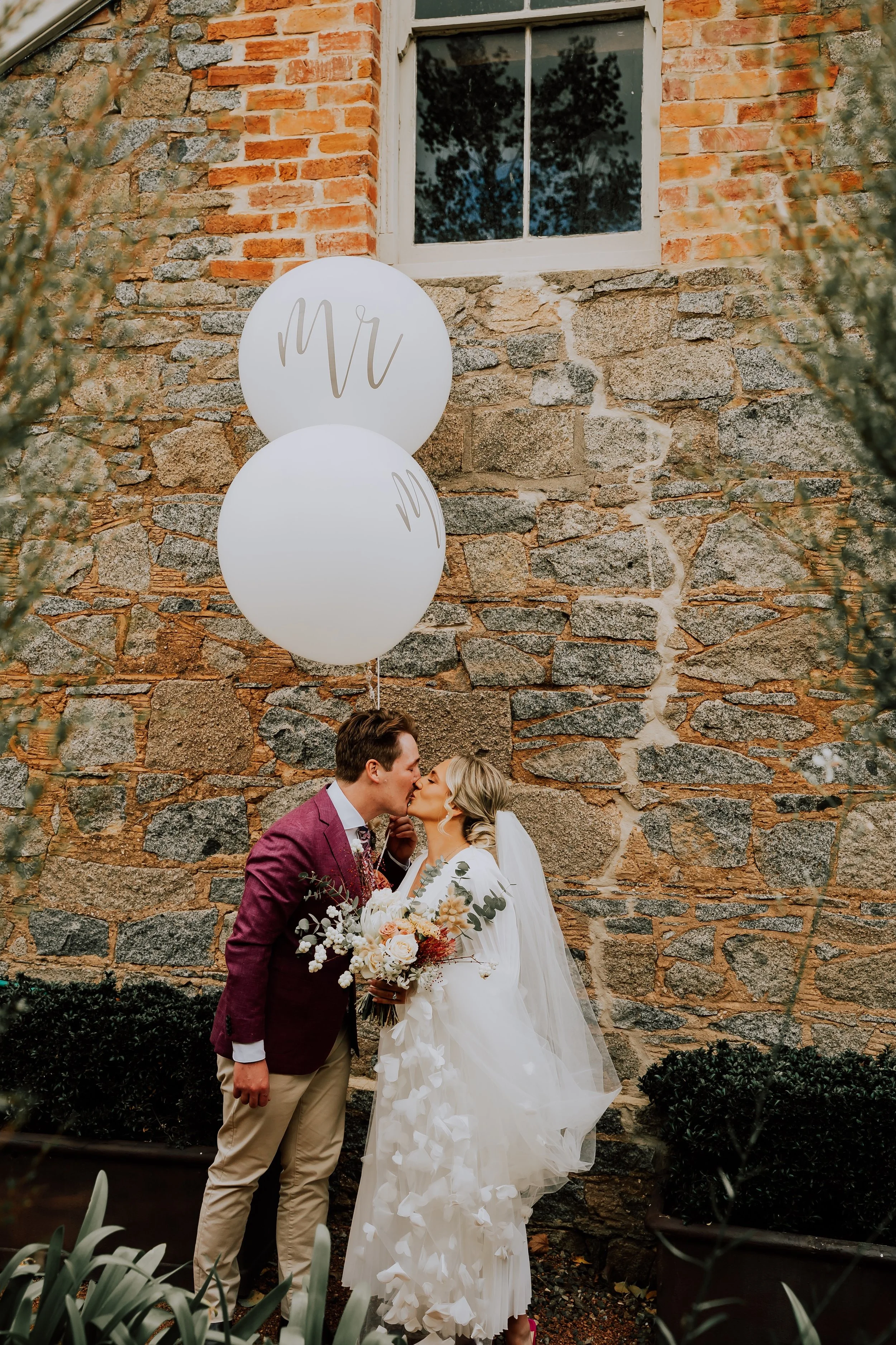 A bride and groom sharing a kiss outdoors against a rustic brick and stone wall. The bride is in a white wedding dress holding a bouquet, and the groom is in a burgundy blazer with beige pants.