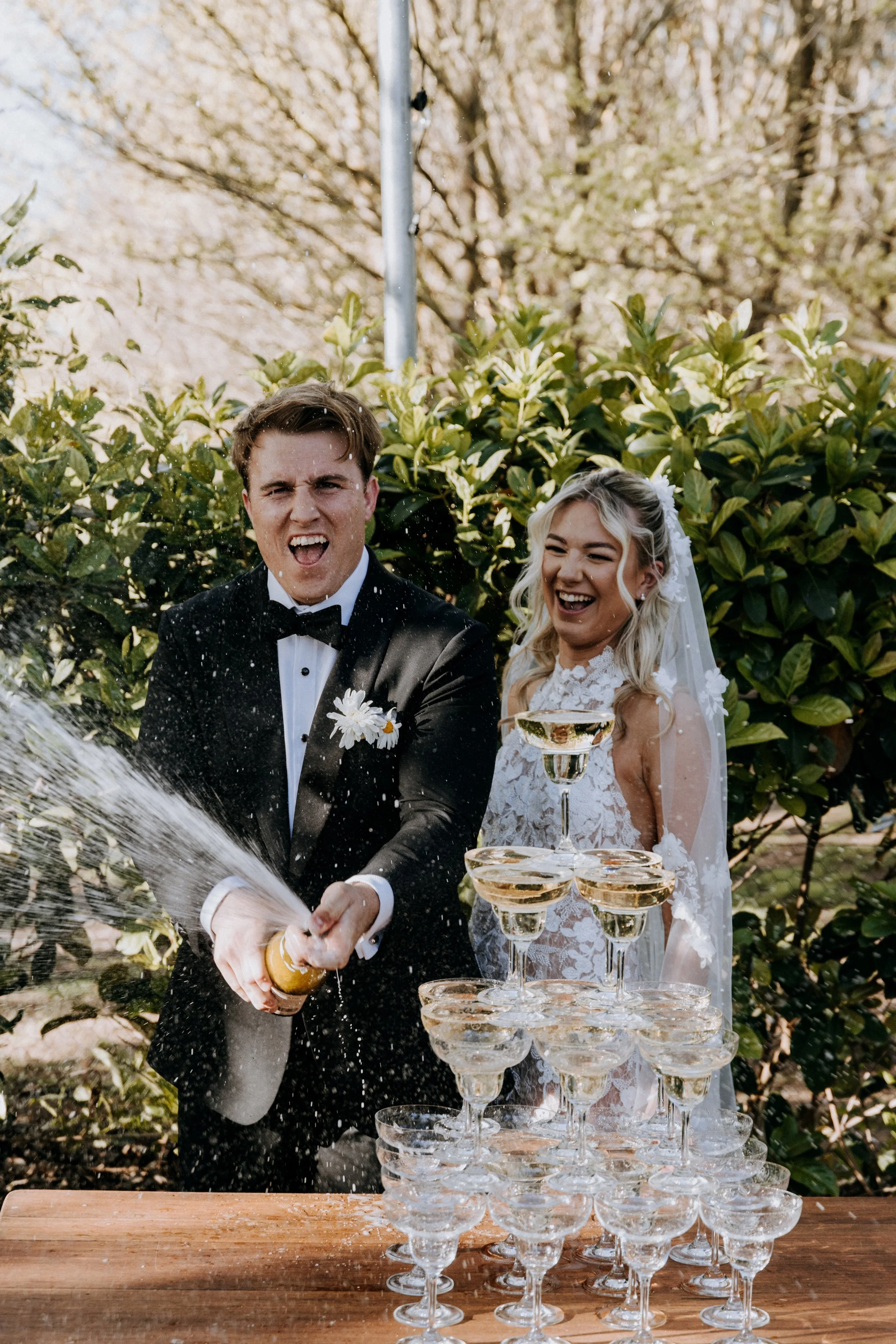 A newlywed couple celebrating by popping champagne, with a pyramid of champagne glasses in front of them, outdoors with greenery and trees in the background.