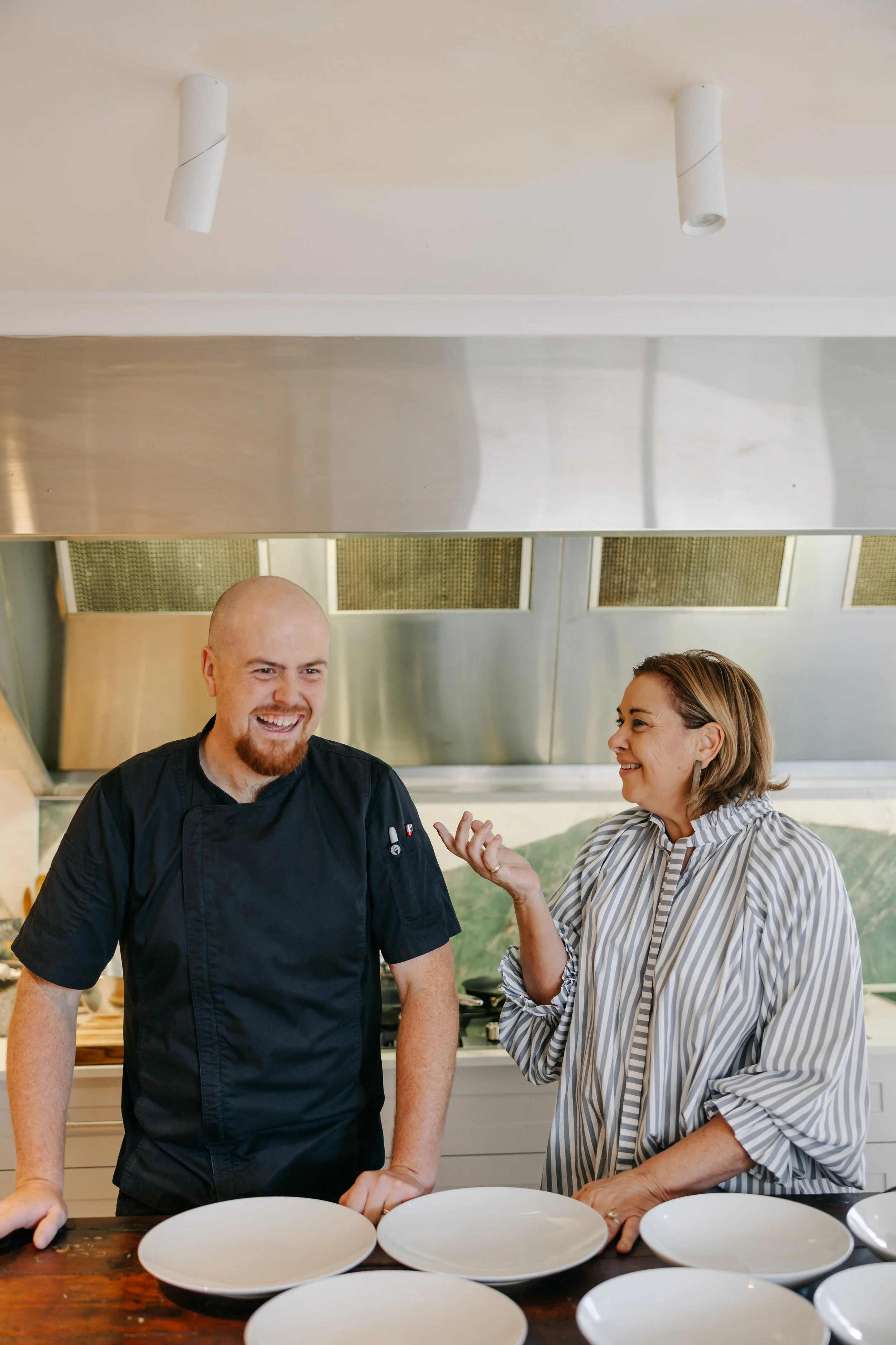 A smiling male chef and a woman with short hair and striped shirt talking in a kitchen with white plates on a wooden table.