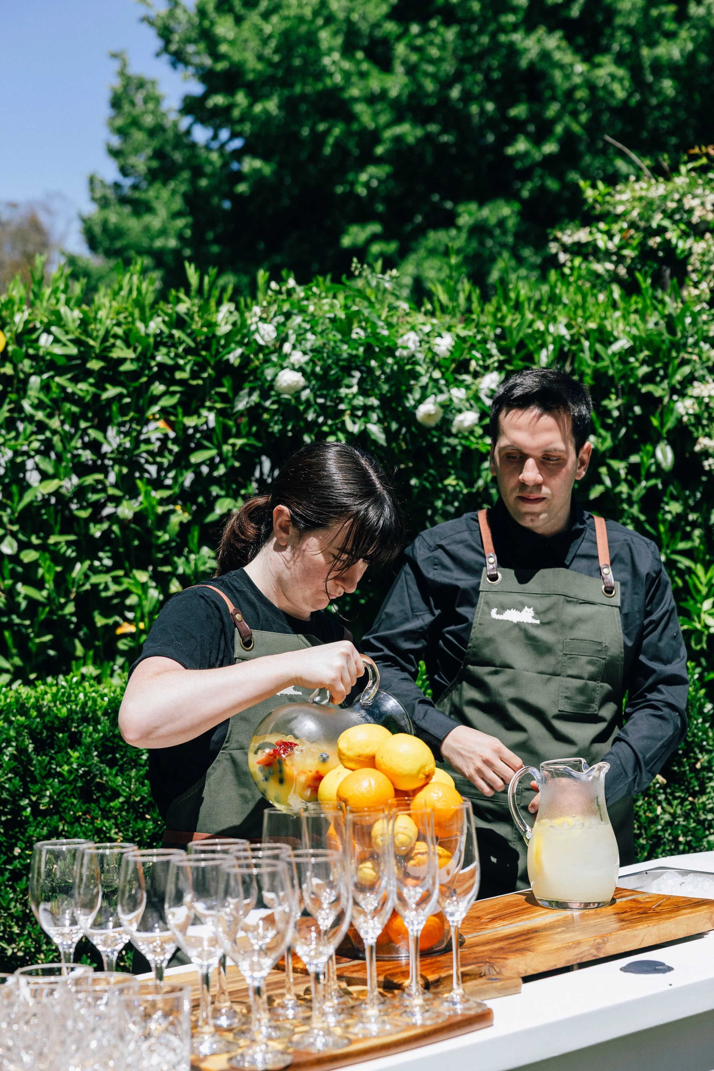 Two people wearing black shirts and green aprons preparing fruit drinks outdoors, with a background of bushes and trees. A glass bowl filled with lemons, oranges, and berries is on the table, along with empty wine glasses lined up on a wooden surface