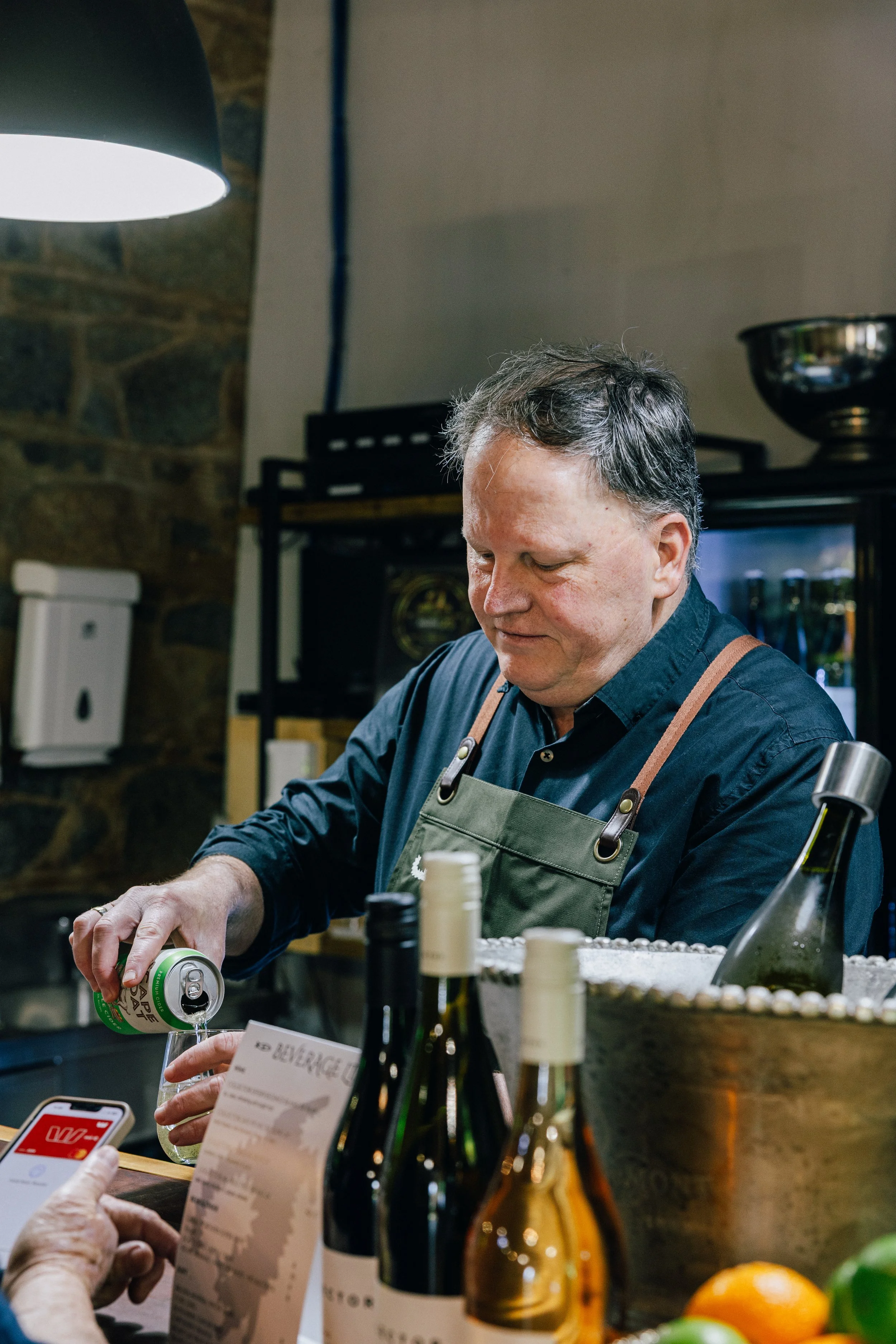 A man with gray hair wearing a black shirt and green apron pouring a canned beverage into a glass at a bar or restaurant. There are wine bottles on the counter in front. In the foreground, a hand holding a phone and a receipt or menu are visible. The
