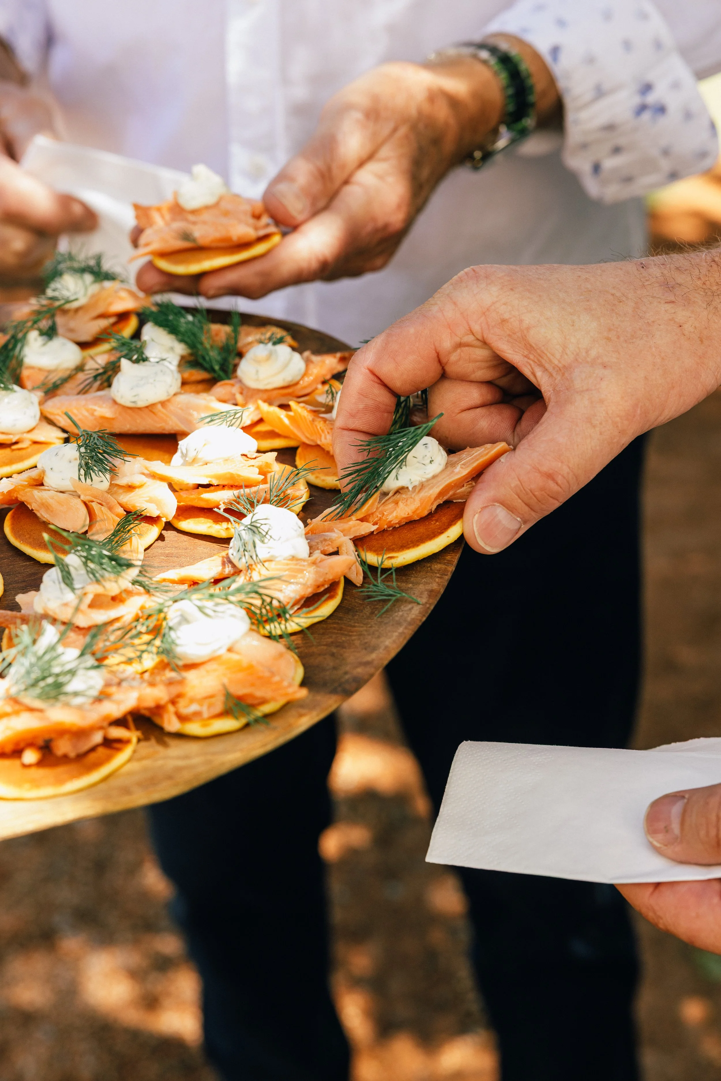 Close-up of a person placing smoked salmon and dill on a plate of canapés, with a hand holding a paper napkin nearby.