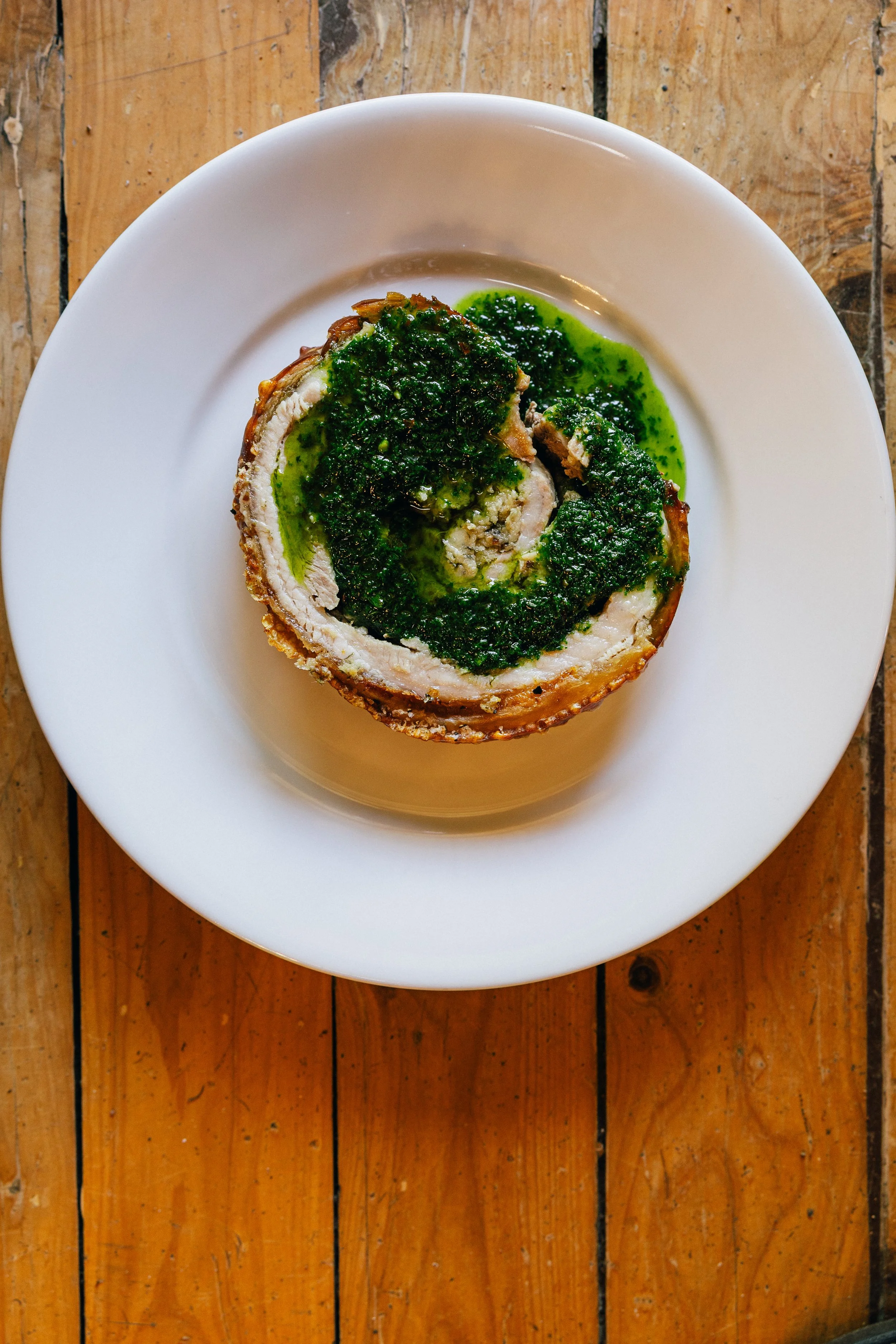 A white plate with a rolled meat dish topped with green herb sauce on a wooden surface.