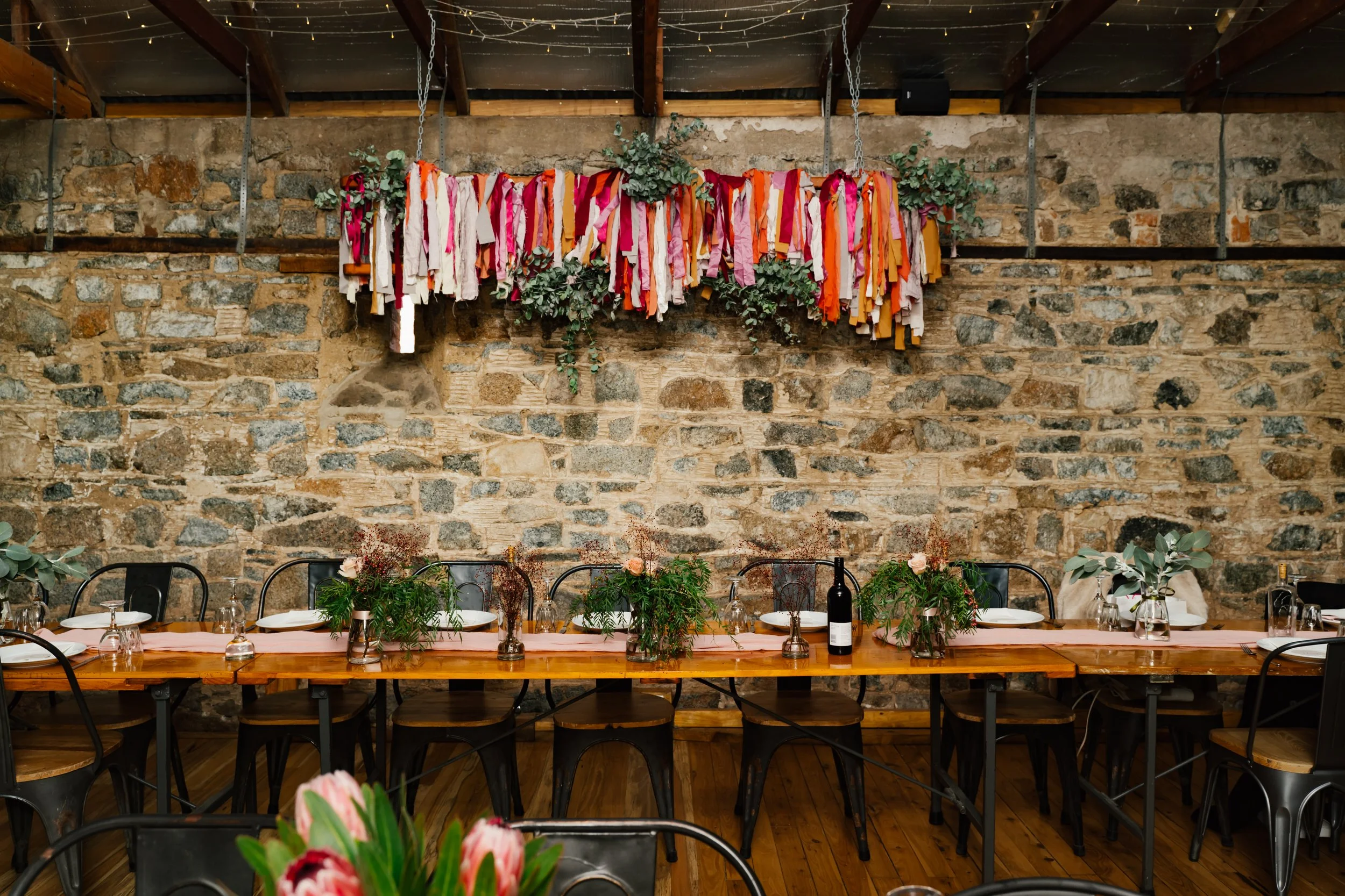 Decorated banquet table in a rustic venue with a stone wall. The table has floral centerpieces and place settings, and a hanging fabric and greenery decoration above.