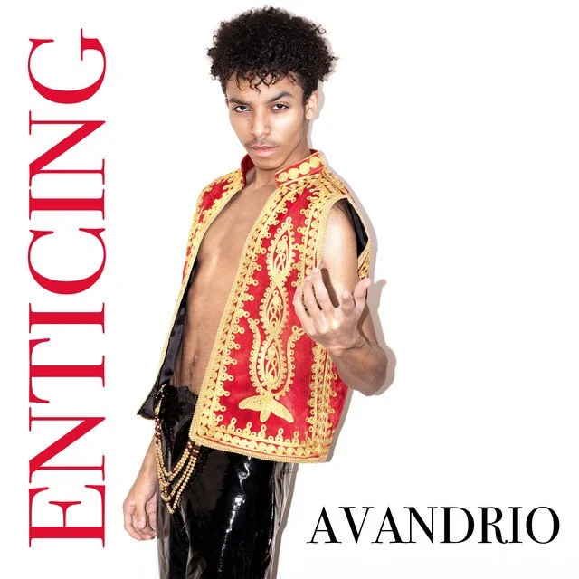 Young man with curly hair wearing a red and gold embroidered vest, black shiny pants, standing against a white background, pointing towards the camera, with the words "ENIGTIC" in large red letters on the left and "AVANDRIO" in black text at the bottom.