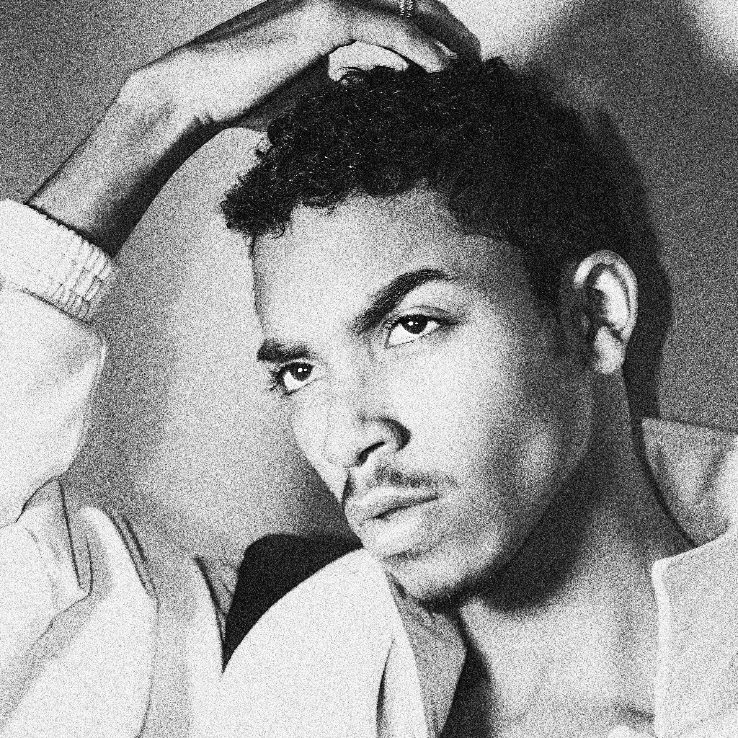 Black and white portrait of a young man with short curly hair, expressive eyes, a slight mustache, and goatee, wearing a white jacket, with his hand resting on his head.