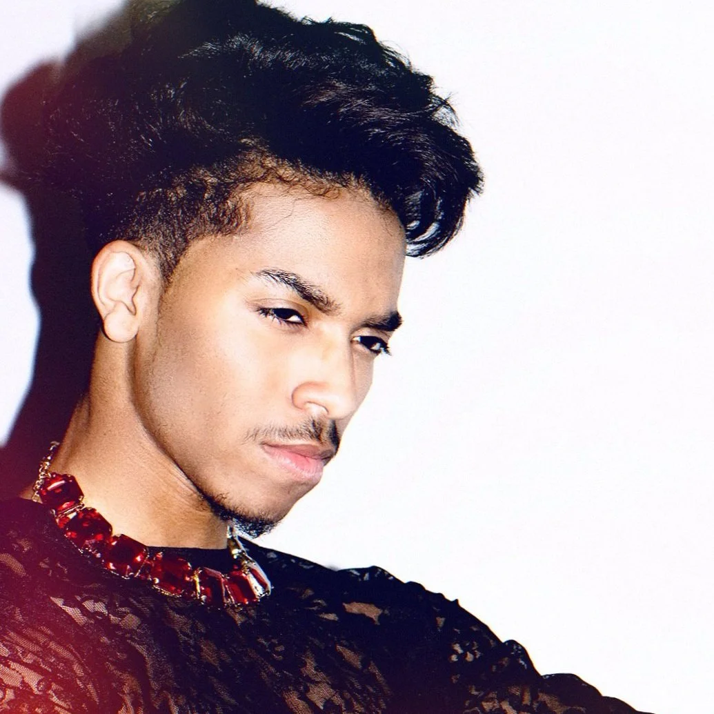 A young man with dark, wavy hair, wearing a black lace top and a red beaded necklace, looking at the camera with a serious expression.