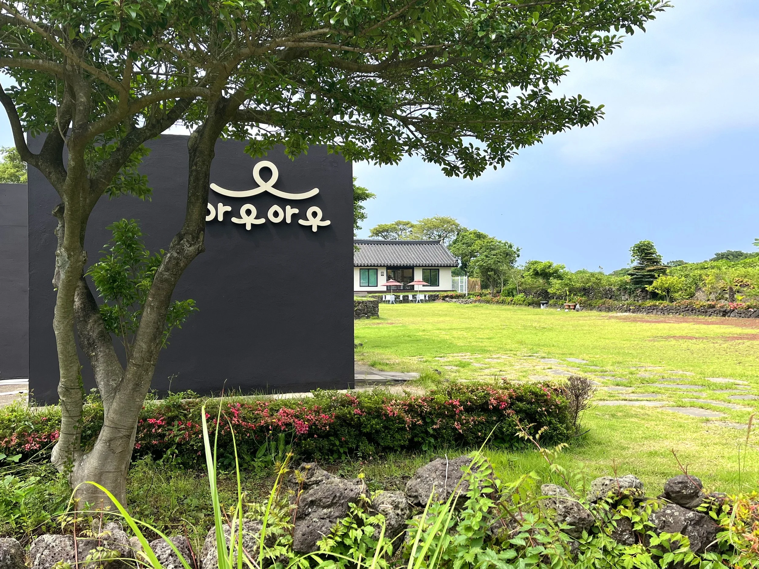 A landscaped garden with greenery, flowers, rocks, and two houses in the background, with a black sign featuring a white logo and text.