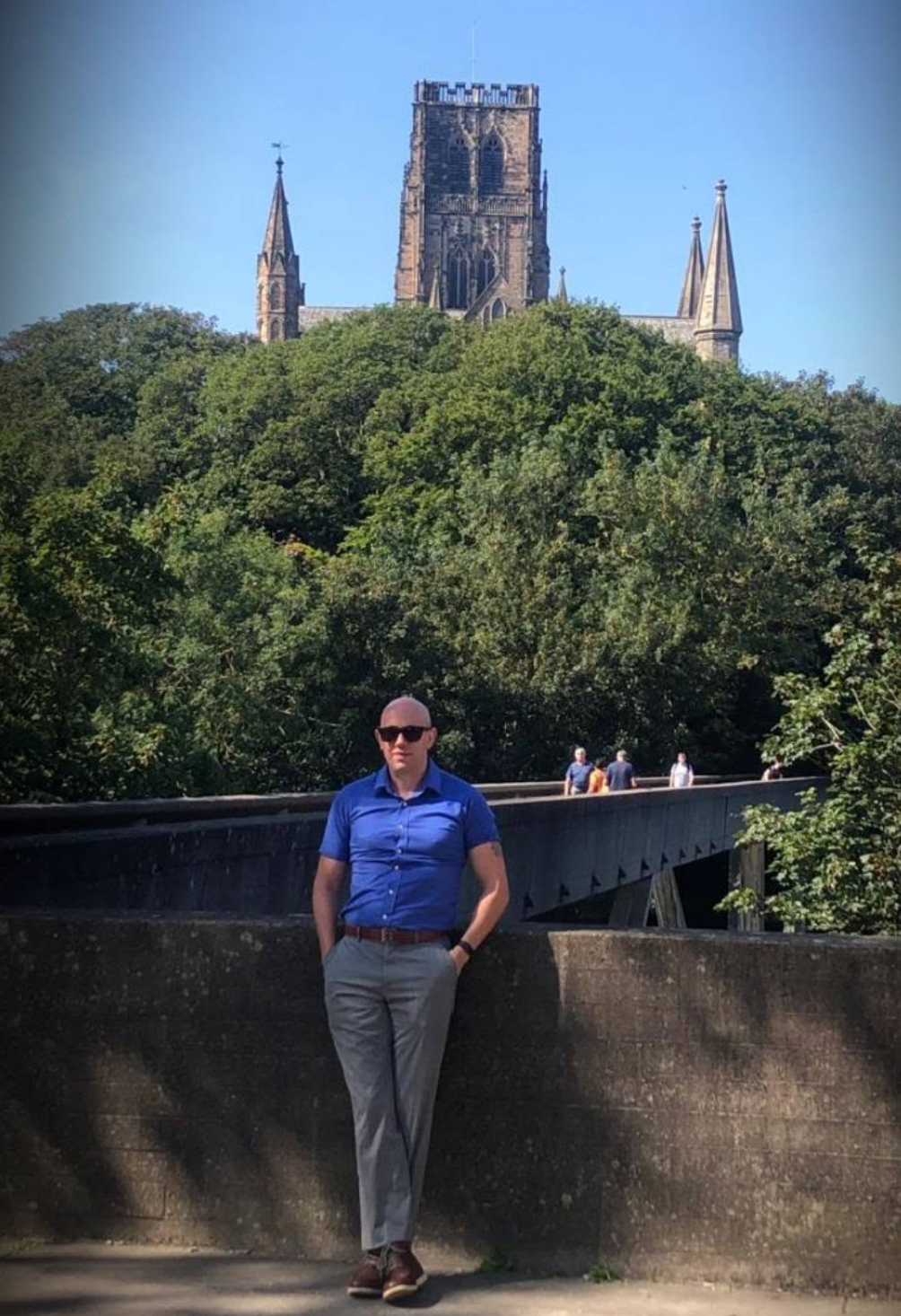 Author and espionage novelist James Burnett standing on a stone bridge, with historic architecture and green trees in the background.