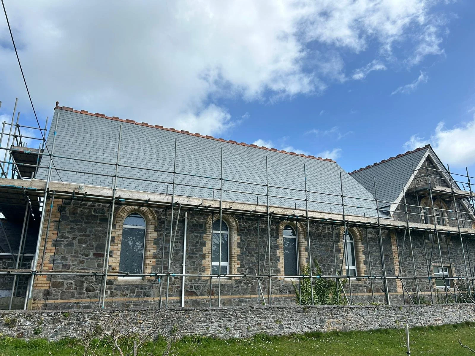 A stone building under construction with scaffolding around it, new roof tiles being installed, and a blue sky with scattered clouds above.