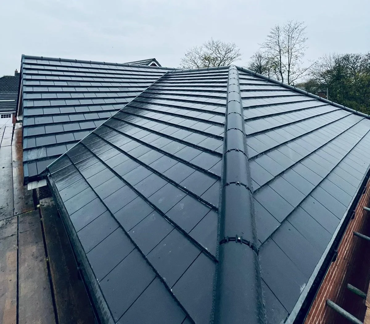 A close-up view of a tiled metal roof on a building with trees and cloudy sky in the background.