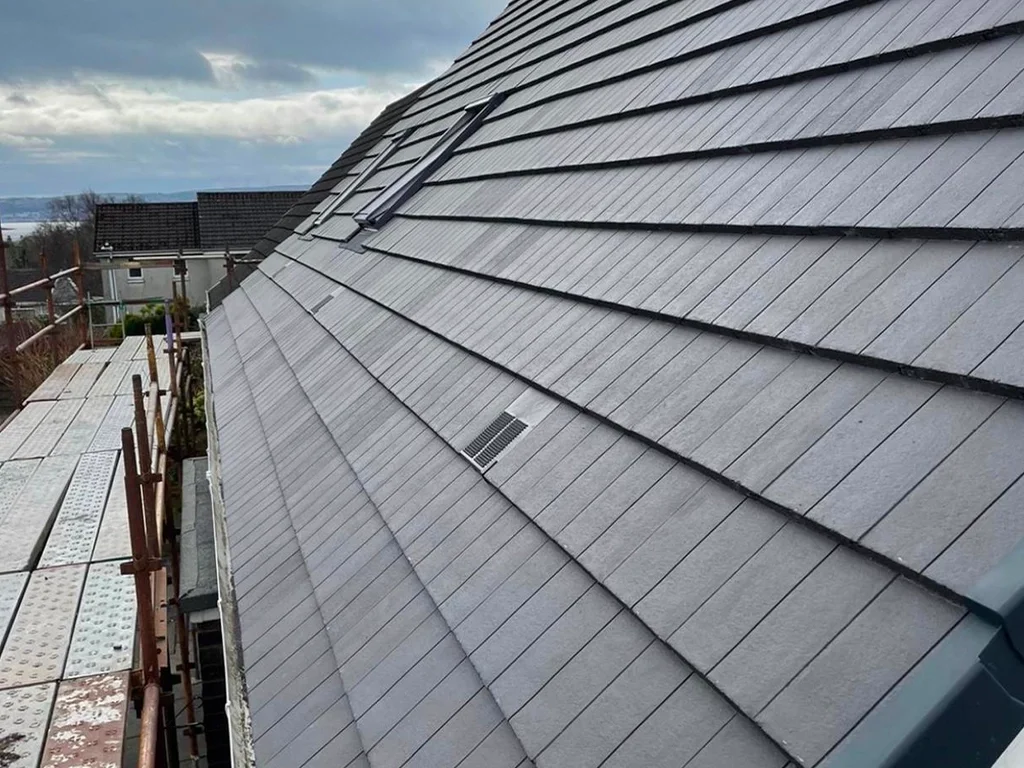 Close-up view of a gray shingle roof with vent openings, some scaffolding and construction materials on the left, and a distant landscape background with cloudy skies.