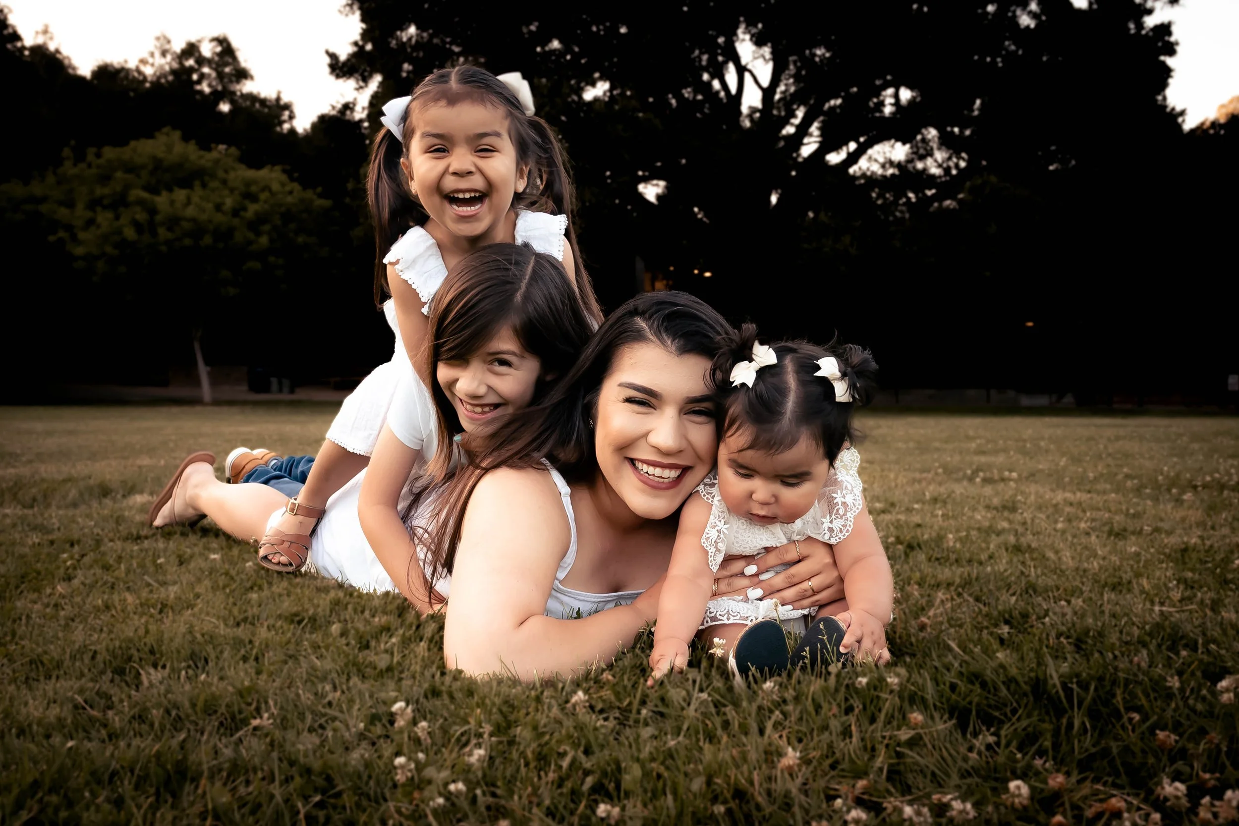 A woman and four young girls lying on the grass in a park, stacking their bodies in a playful and joyful manner during sunset.