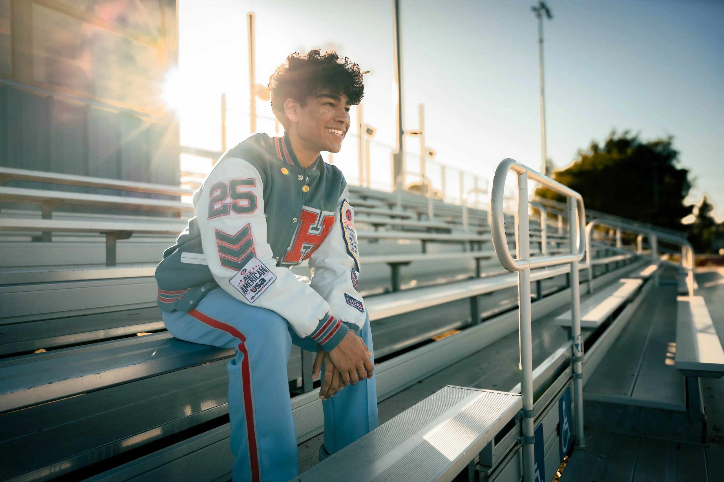 Young man in varsity jacket sitting on bleachers at an outdoor stadium, smiling, with sunlight behind him.