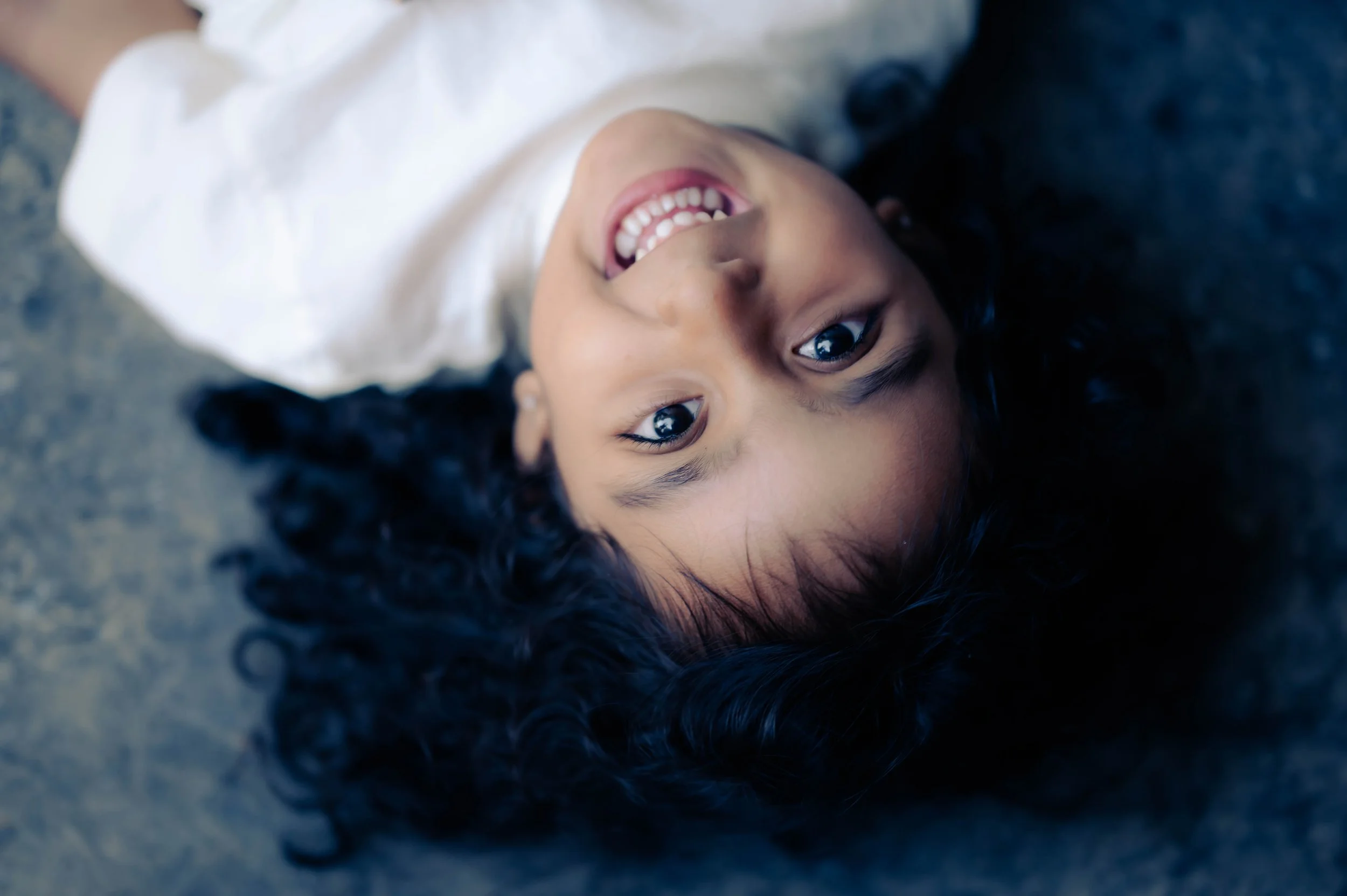 A young girl with curly black hair lying on her back on a dark surface, smiling and looking up at the camera.