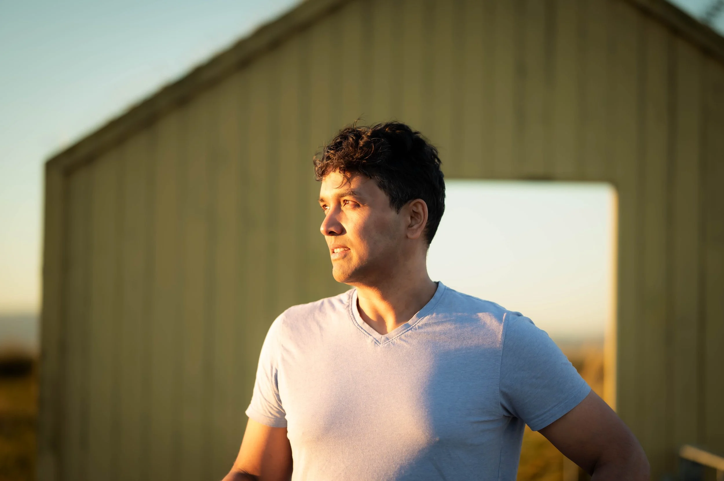 A young man standing outdoors during sunset near a light yellow barn with side opening.
