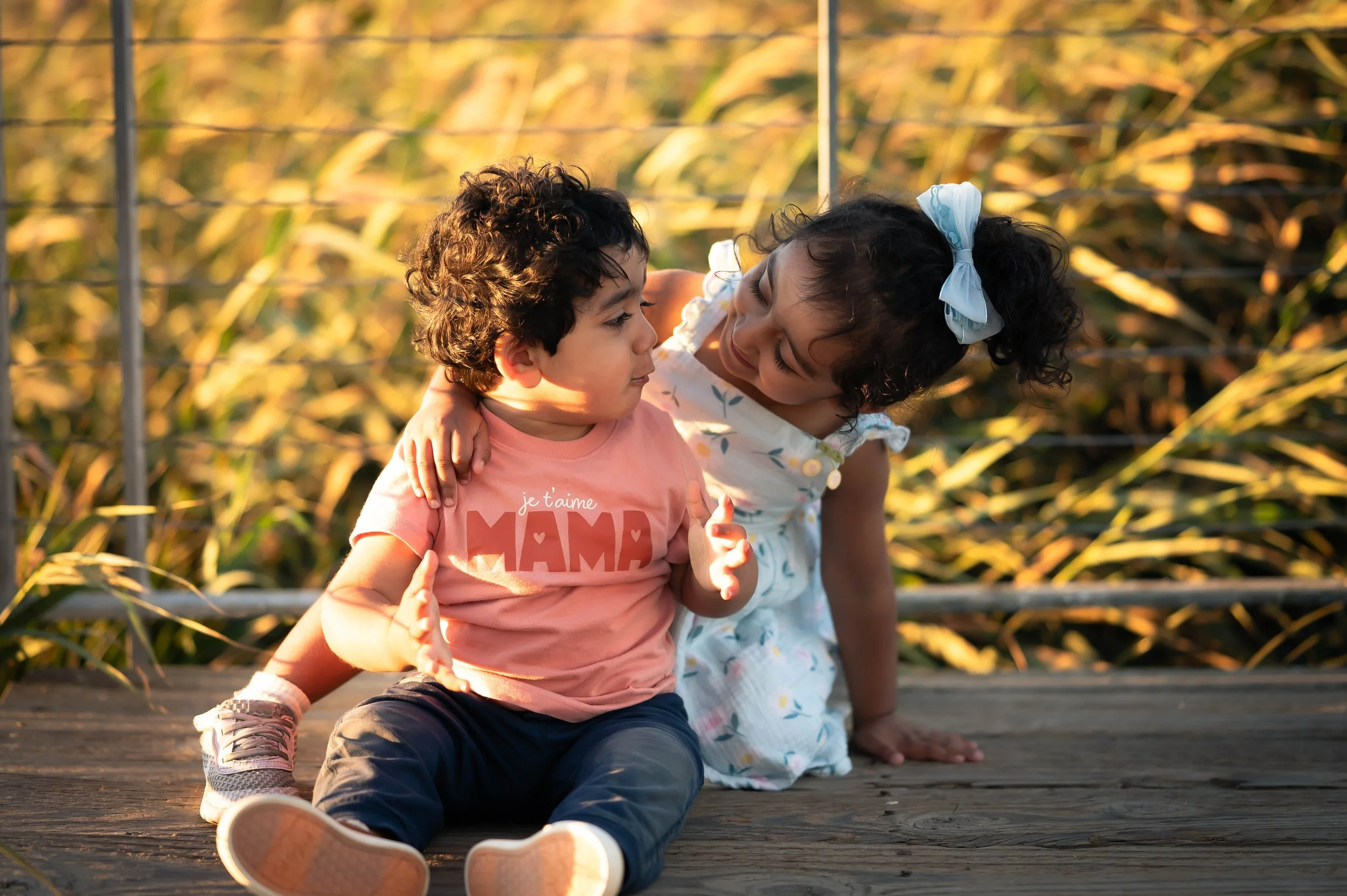A young boy and girl sitting on a wooden bridge, with the girl leaning in to look at the boy. The boy wears a pink T-shirt with mairie and the girl wears a white dress with colorful patterns. Sunlight filters through the background foliage.