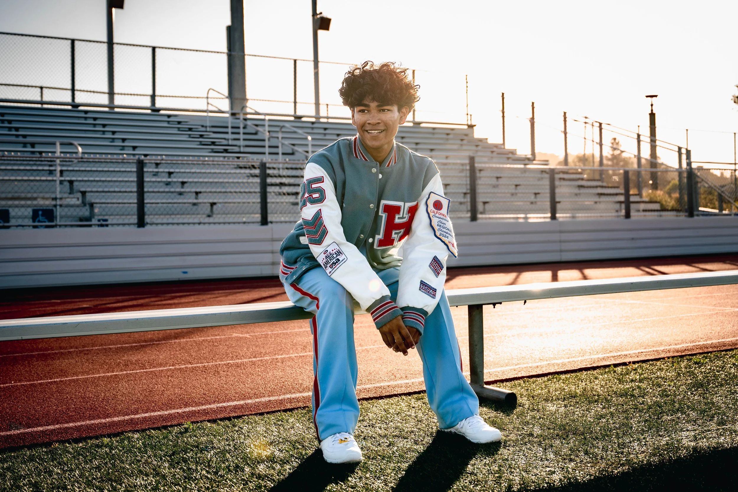 Young person sitting on a stadium bench on a running track, wearing a varsity jacket with patches, smiling, during sunset.