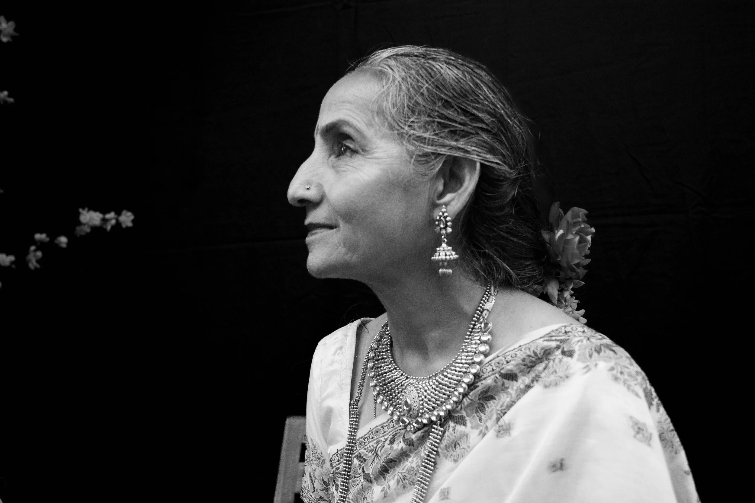 Black and white photo of an elderly woman in traditional attire with jewelry and flowers in her hair, facing left.