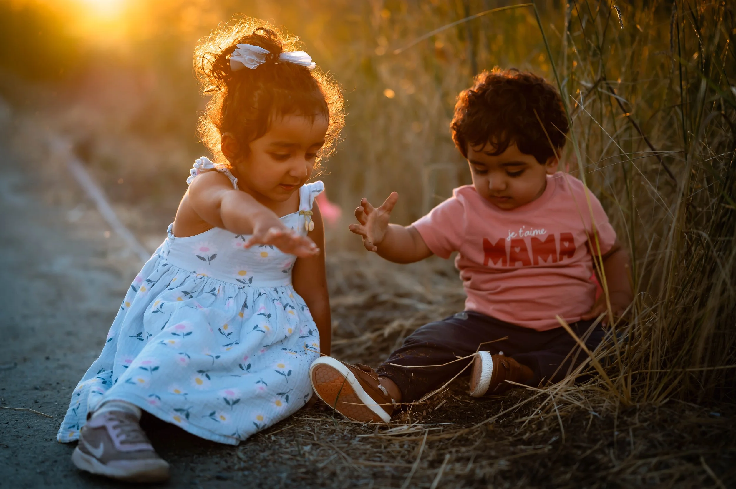 Two young children, a girl in a blue dress and a boy in a pink t-shirt, playing outdoors during sunset among tall grass.