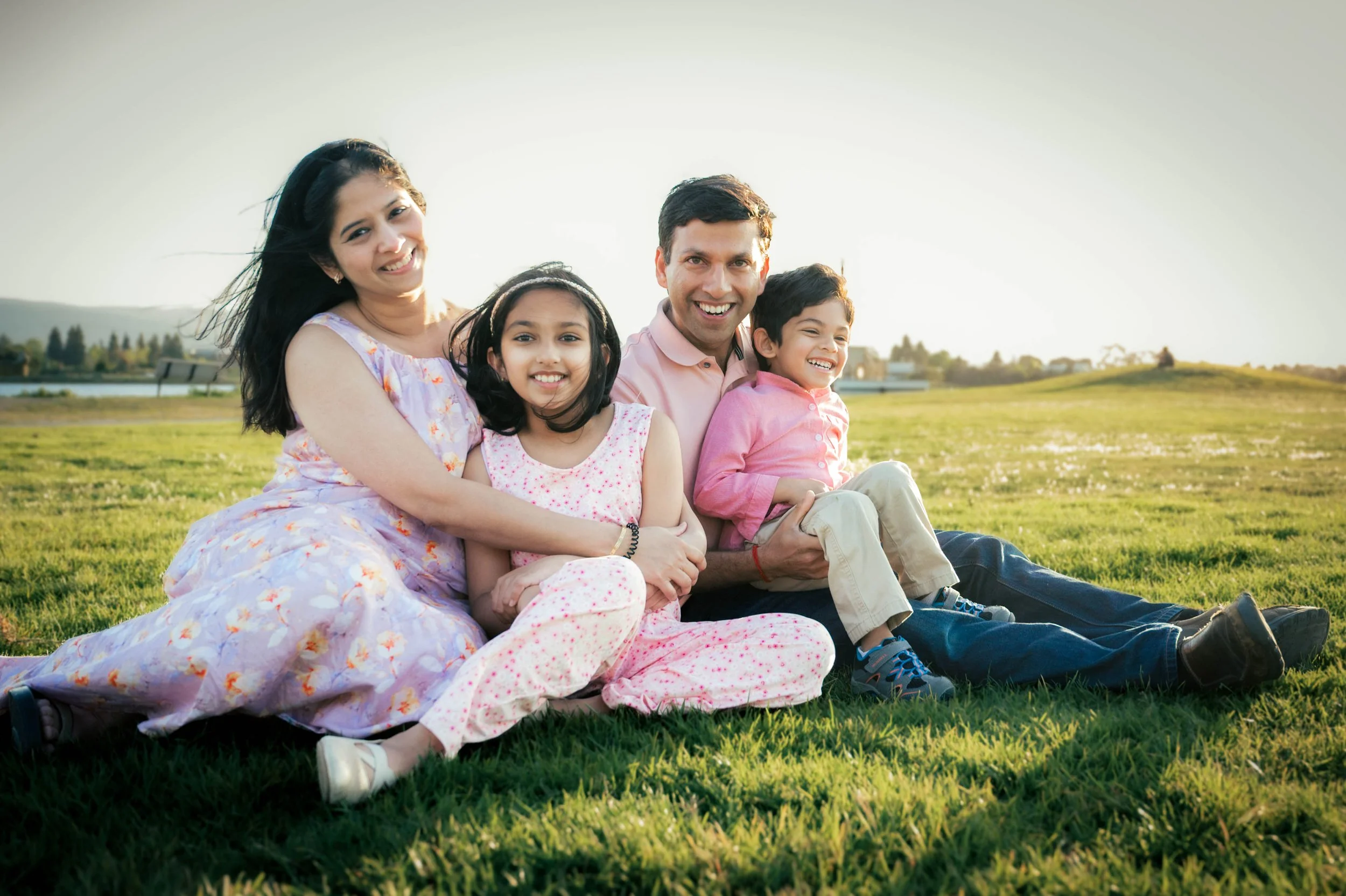 A family of four sitting together on a grassy field outdoors, all smiling and enjoying the sunlight on a clear day.
