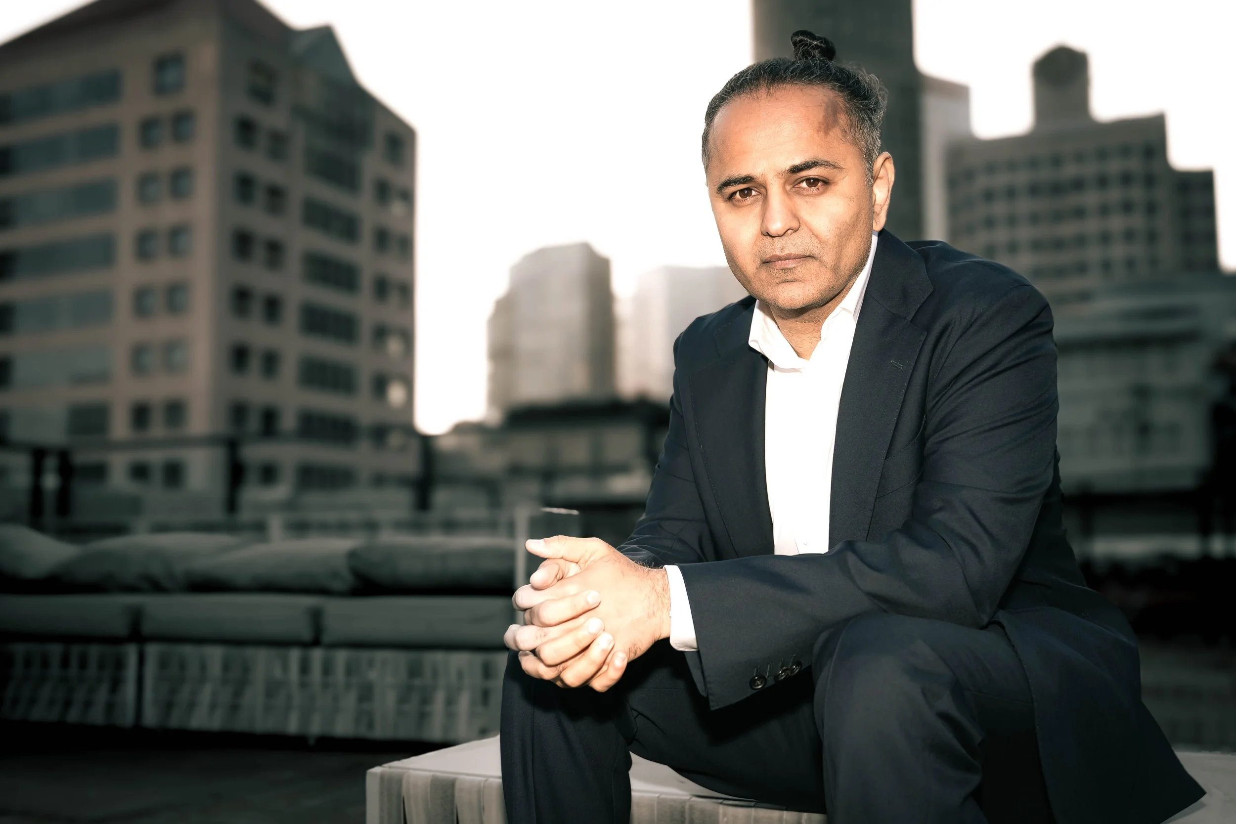 A man in a black suit sitting on a rooftop with city buildings in the background during daylight.