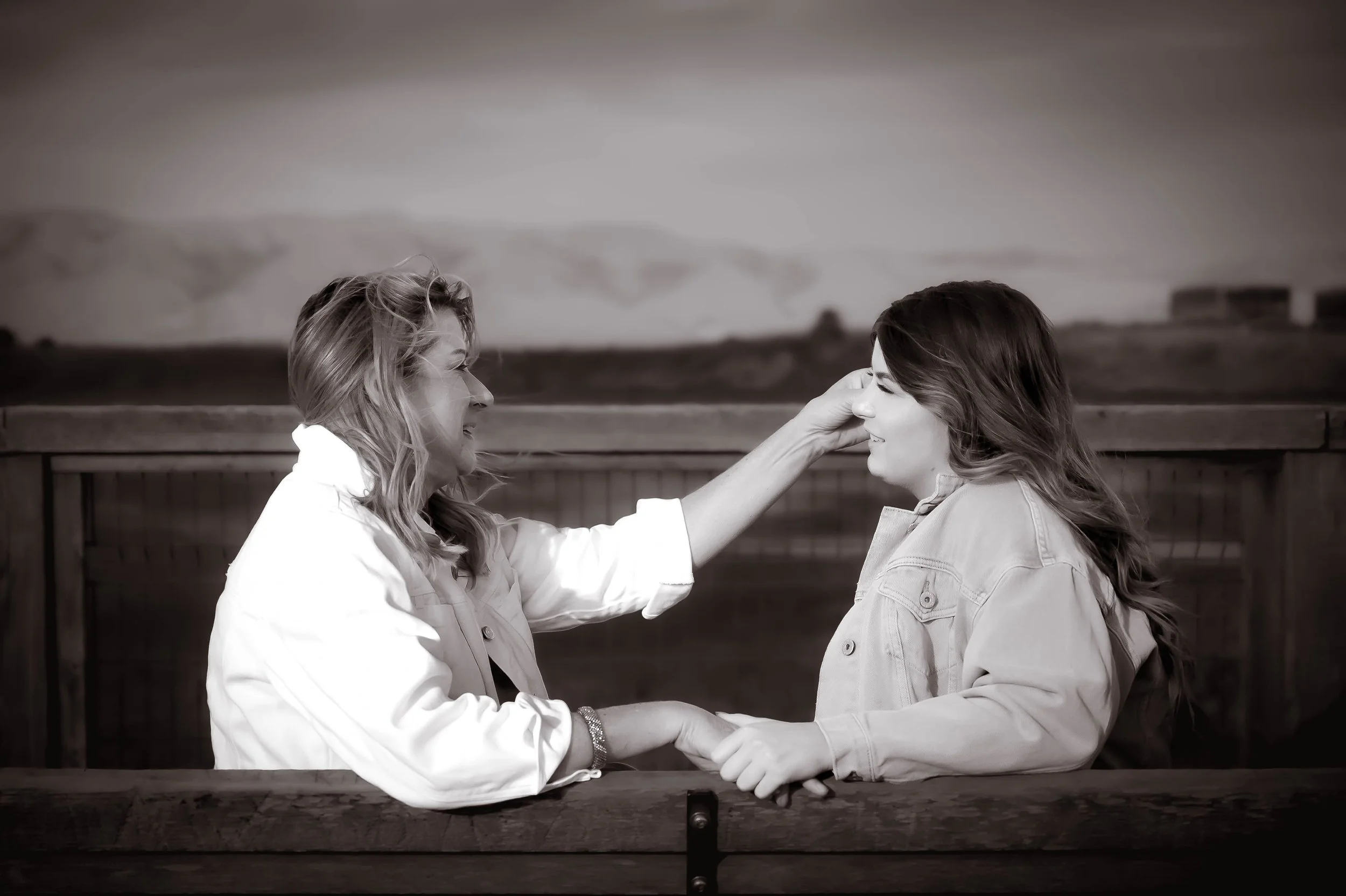 Two women sharing a tender moment on a wooden bench, with one gently touching the other's face and holding hands, outdoors with a blurred landscape in the background.