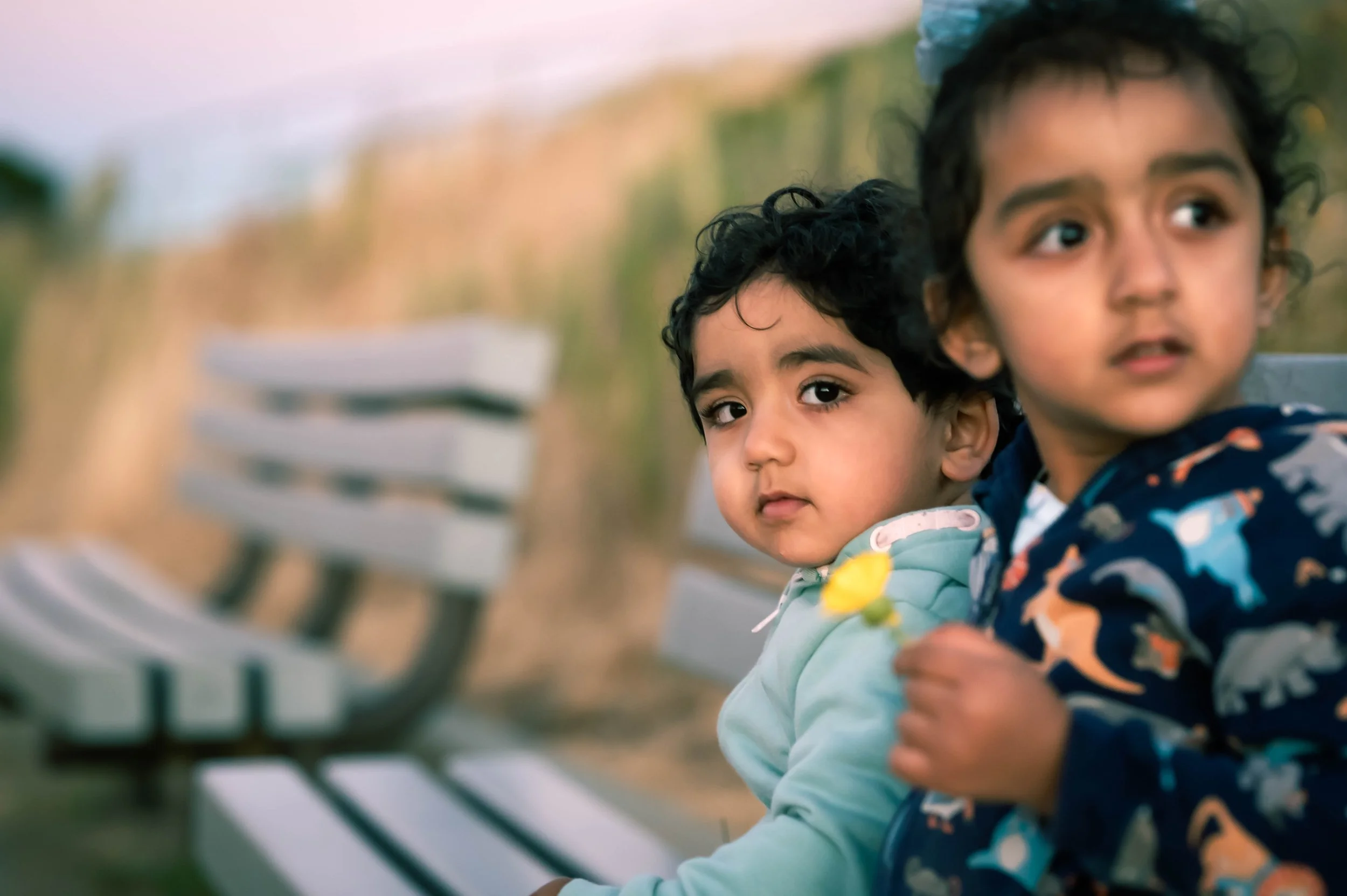 Two young children with dark curly hair sitting on a park bench, with one child holding a yellow flower. They are outdoors, with a blurred background of a natural setting.